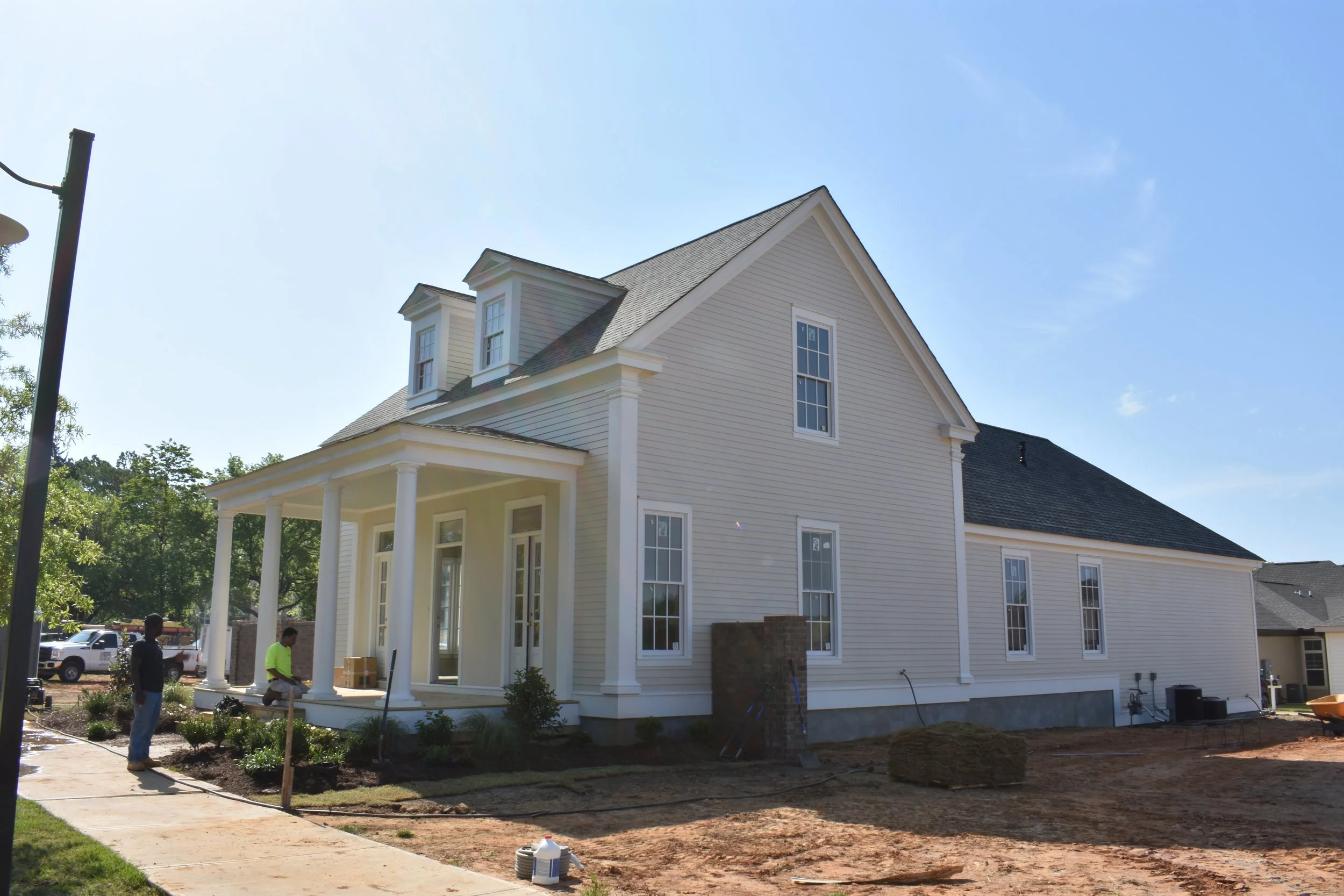 A white house under construction with a front porch supported by white columns, multiple windows, and a dark roof. Two workers are present, one standing and another kneeling, on a construction site with dirt and construction materials visible in the 