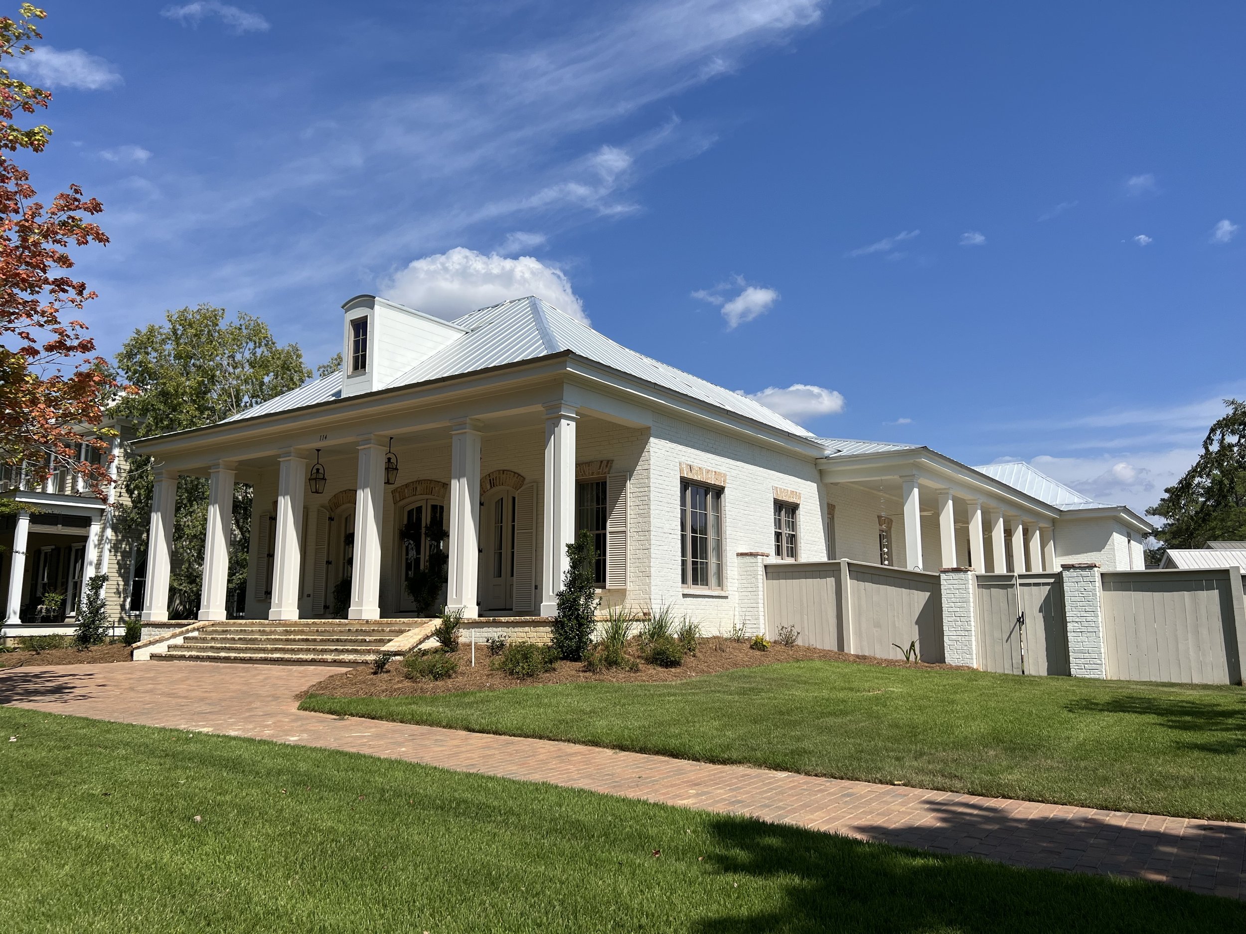 A large, white, historic-style house with a metal roof, front porch with columns, brick stairs, and a well-manicured lawn under a bright blue sky with some clouds.