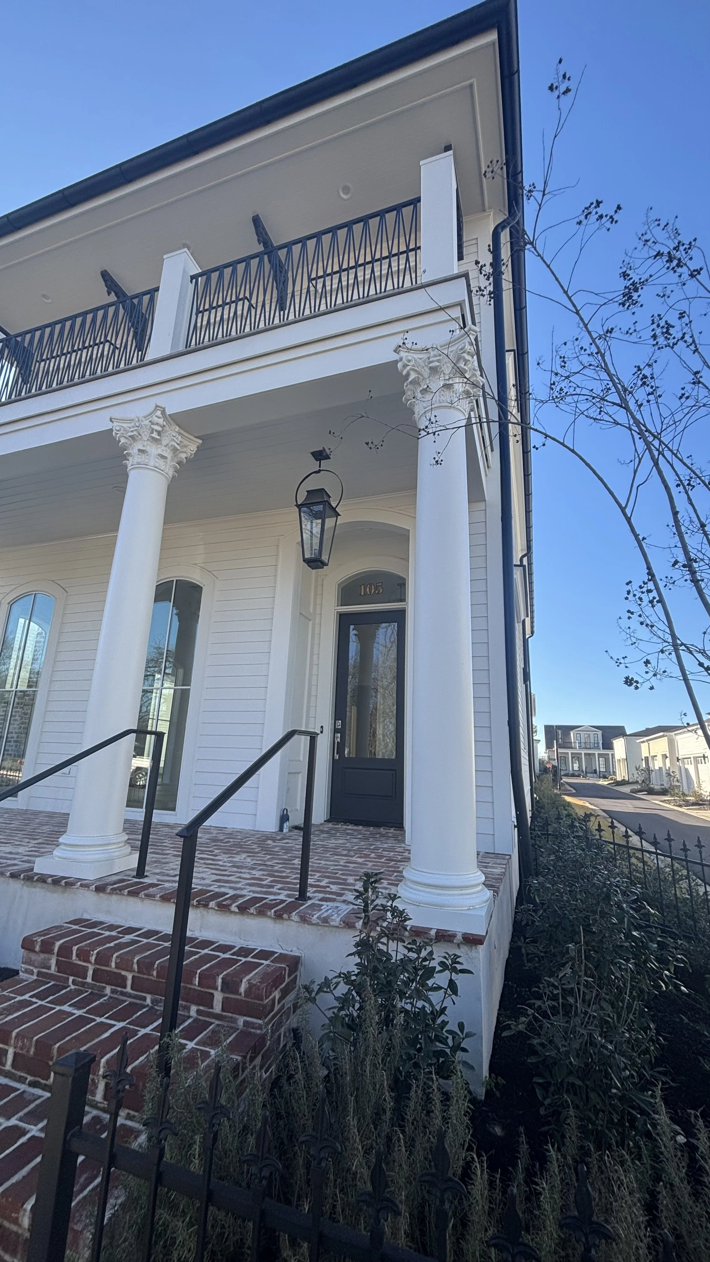 A two-story white house with a covered porch, brick steps, black handrails, and tall white columns with classical capitals. There is a black front door with the number 105 above it, a hanging lantern, and a balcony with black railing above the porch.