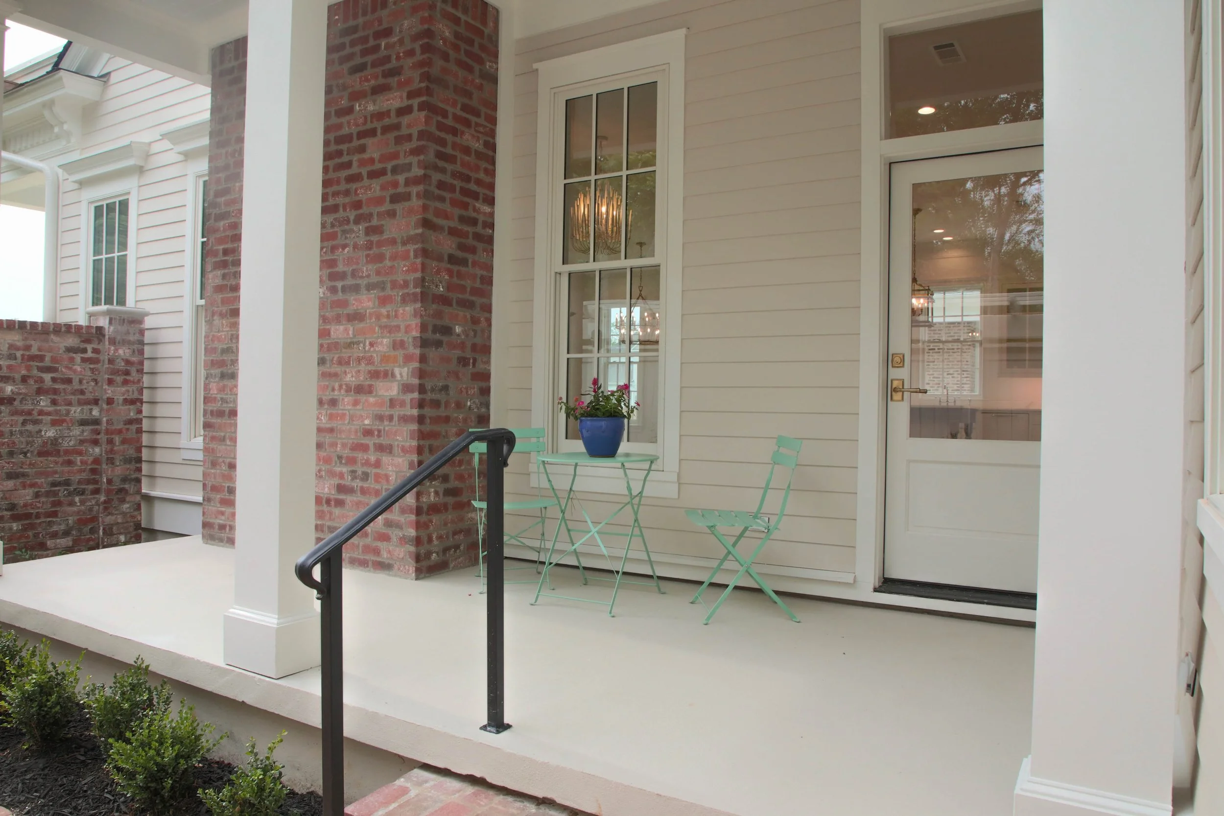 A porch with a small teal table and two teal chairs, one with a flowerpot containing pink flowers on the table, located in front of a house with beige siding and a brick column.