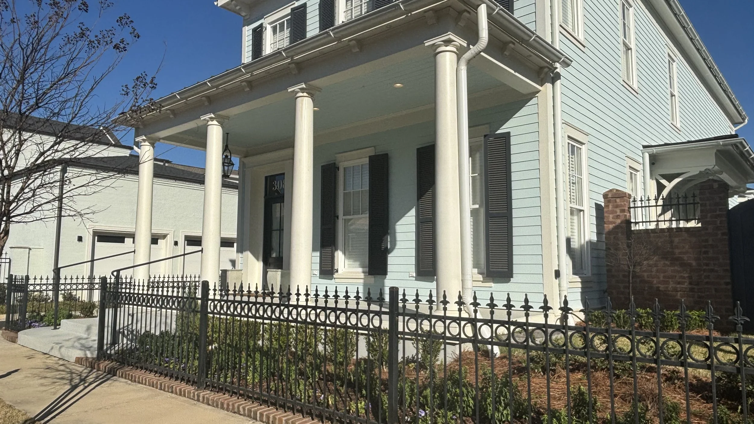 Front view of a light blue two-story house with black shutters, white columns, and a small front garden enclosed by a black metal fence.