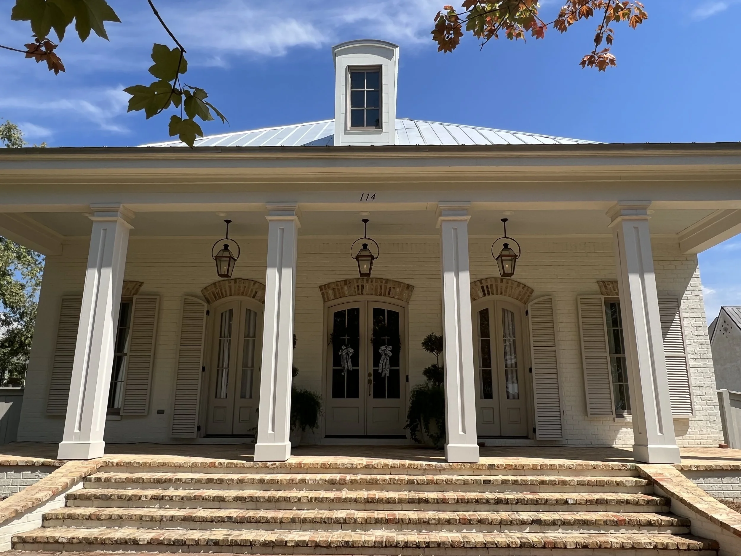 Front view of a white house with brick steps leading to the front door, four pillars supporting the porch roof, and a metal roof with a small dormer window. There are trees with leaves in the foreground and a clear blue sky.