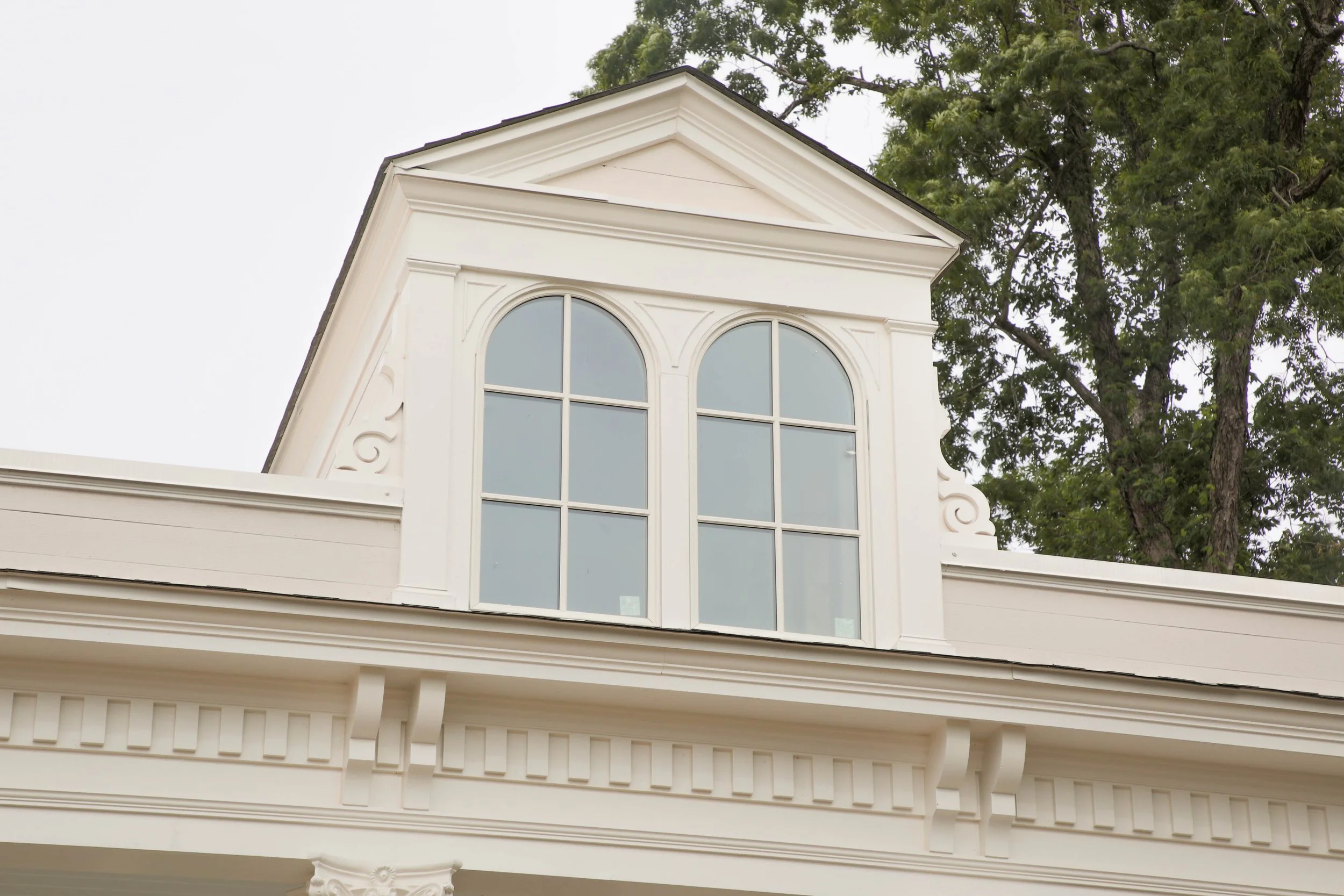 Close-up of a white house with decorative architectural details, featuring large arched windows, part of the roof, and a tree in the background.