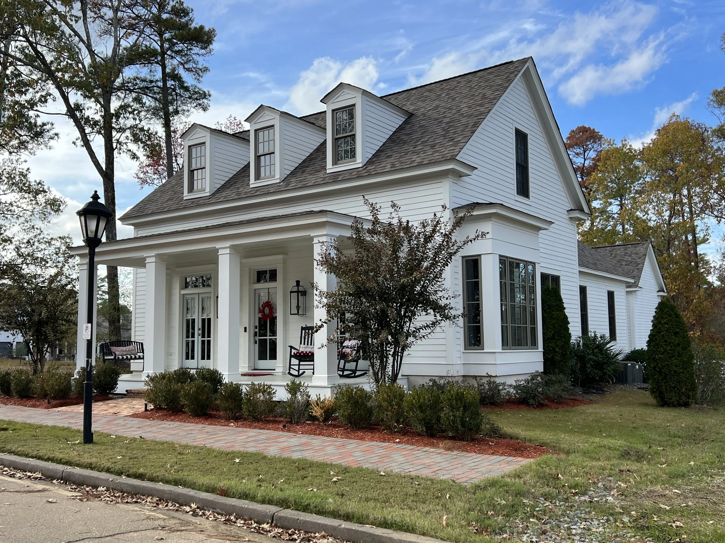 White two-story house with a front porch, black rocking chairs, and a hanging wreath on the door, surrounded by a landscaped yard with bushes and trees, under a partly cloudy sky.