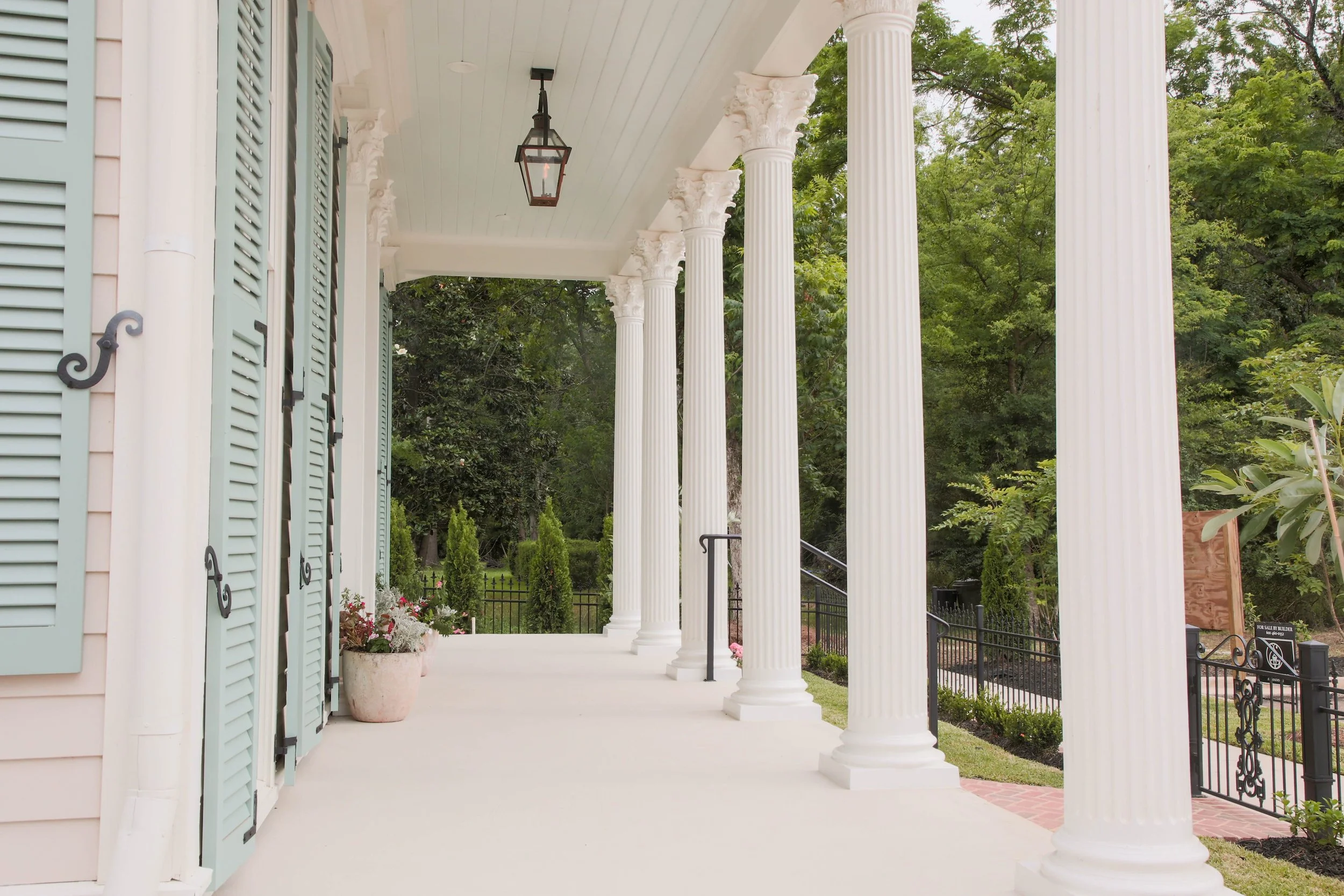 Front porch of a house with white columns and pastel green shutters, with a hanging lantern and potted plants, surrounded by greenery.