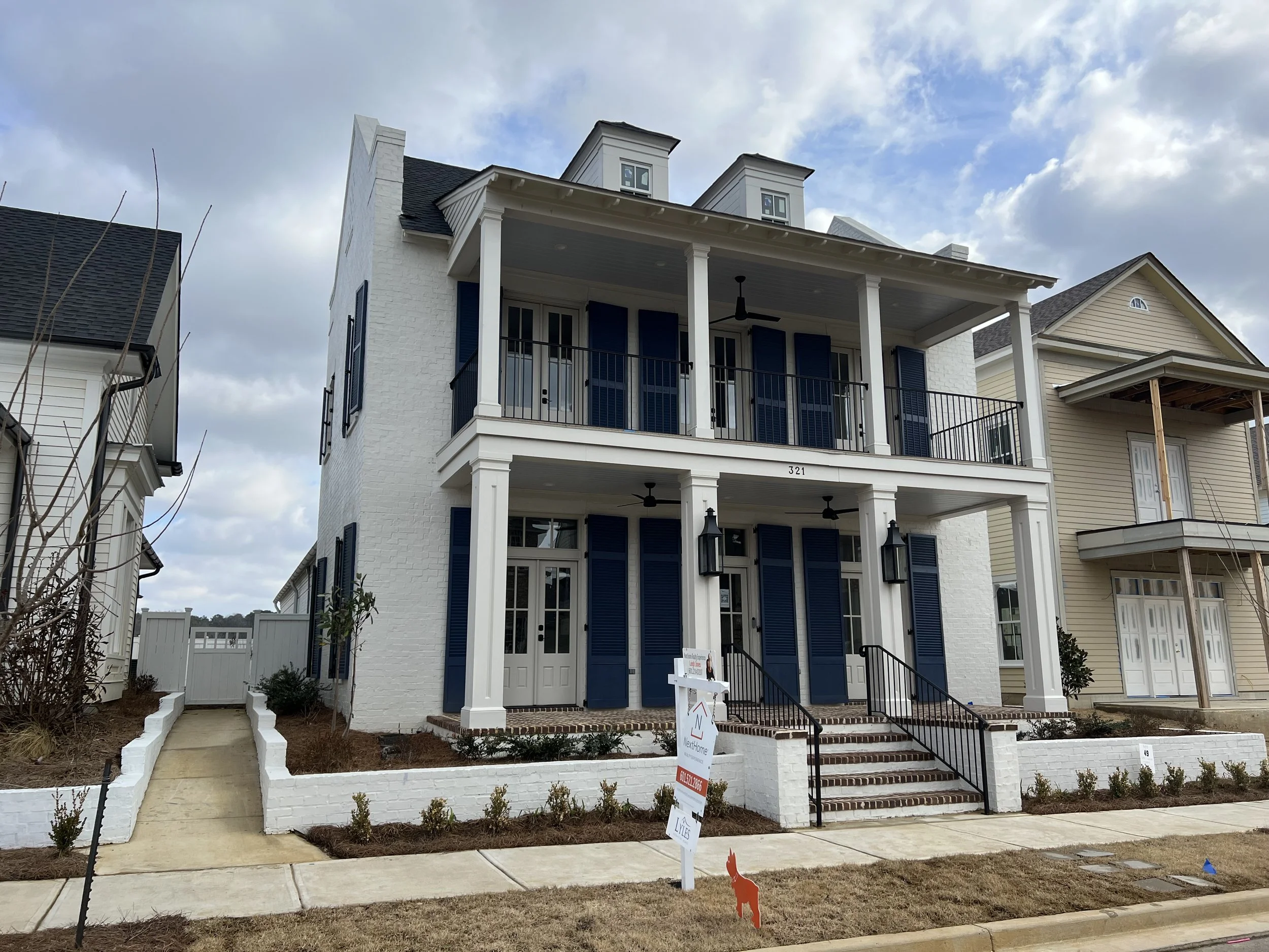 A newly built, three-story house with a white brick exterior, blue shutters, and two front balconies with black railings. The house has a small front porch with stairs and large windows. There is a sidewalk and a small garden in front, with a sign on