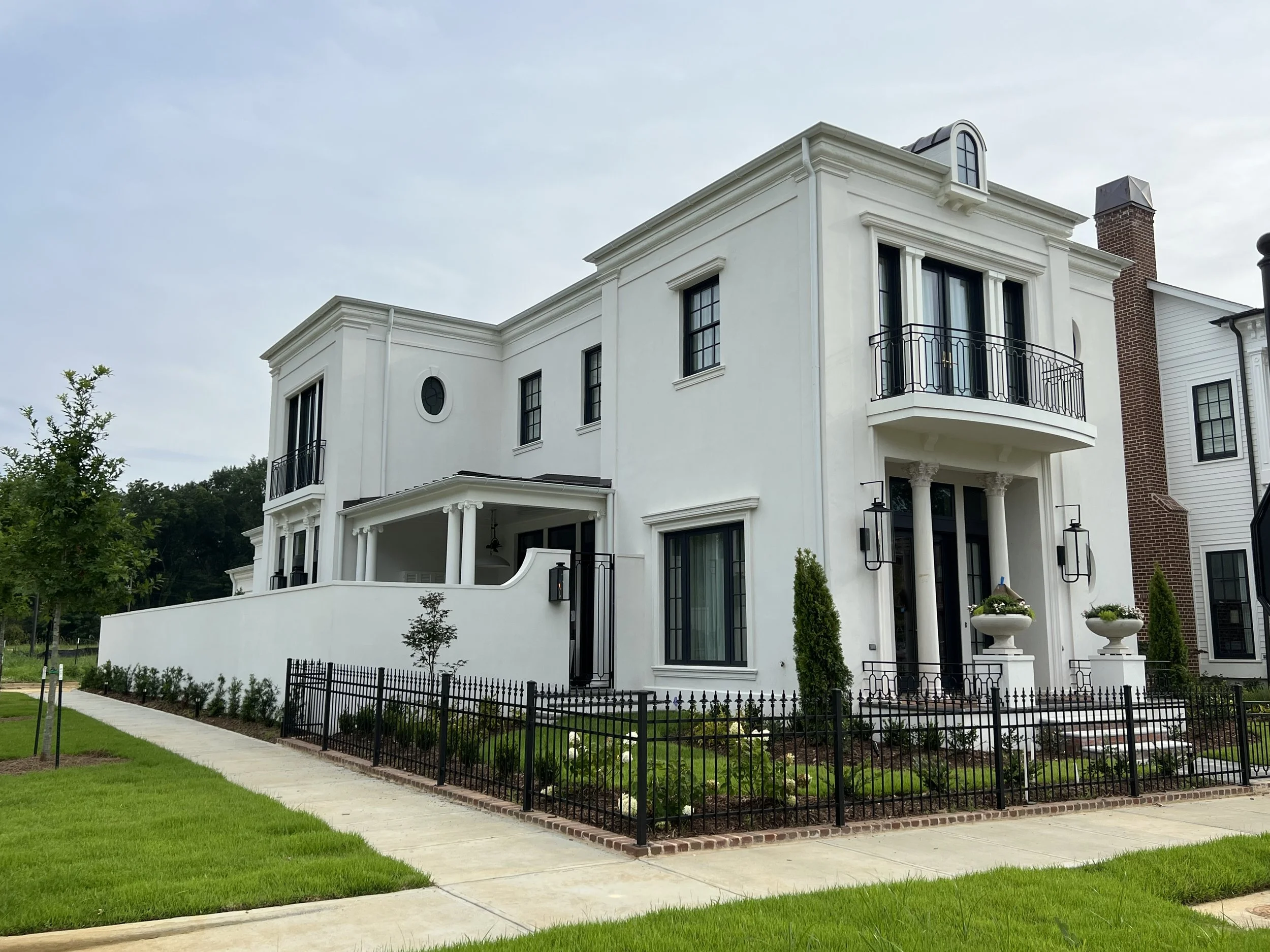 A white three-story house with black accents, a small balcony on the second floor, and a front porch with columns, surrounded by a black fence and landscaped yard.