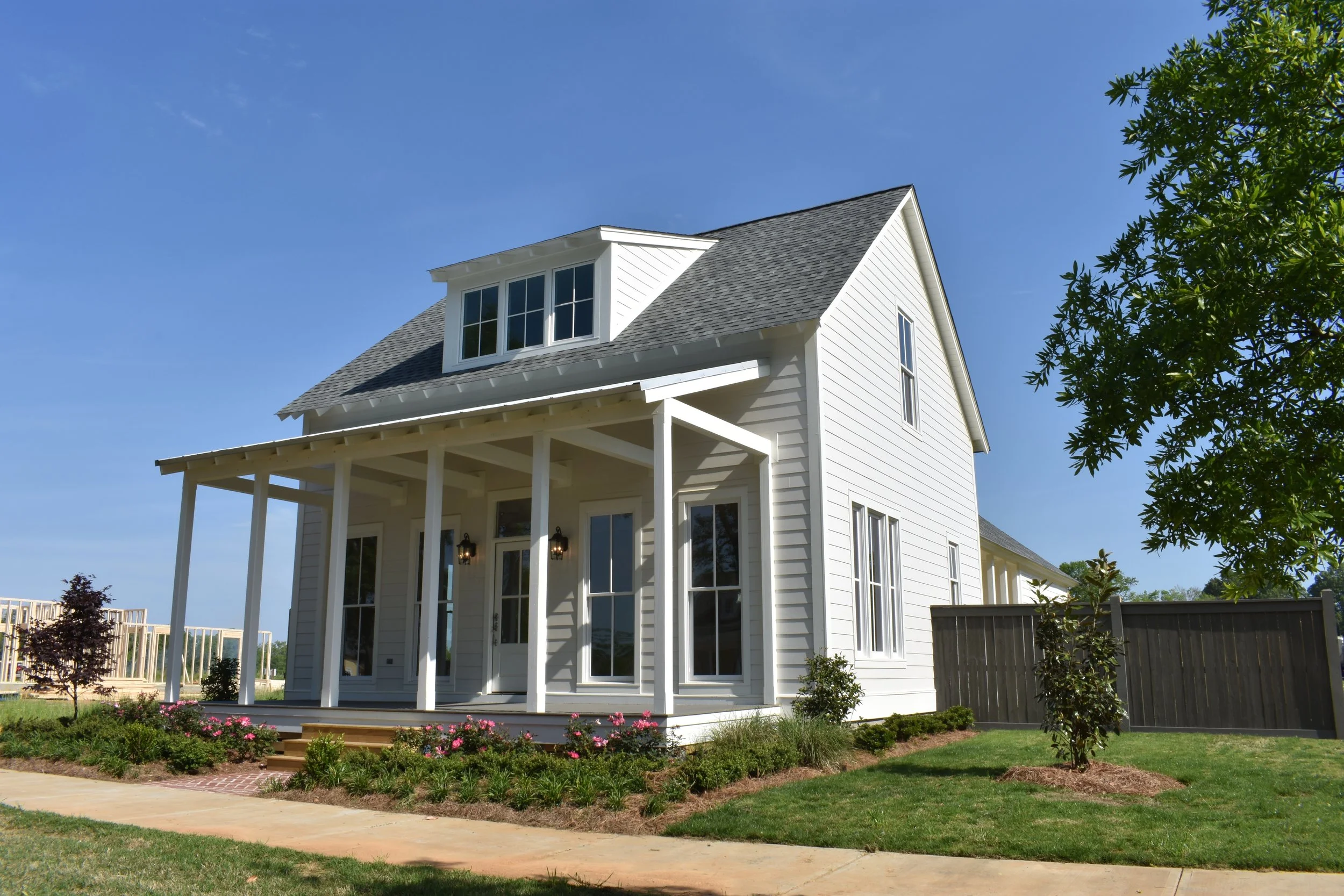 A white, two-story house with a front porch, multiple windows, and a gable roof, set in a landscaped yard with green grass, small trees, and flowering bushes under a blue sky.