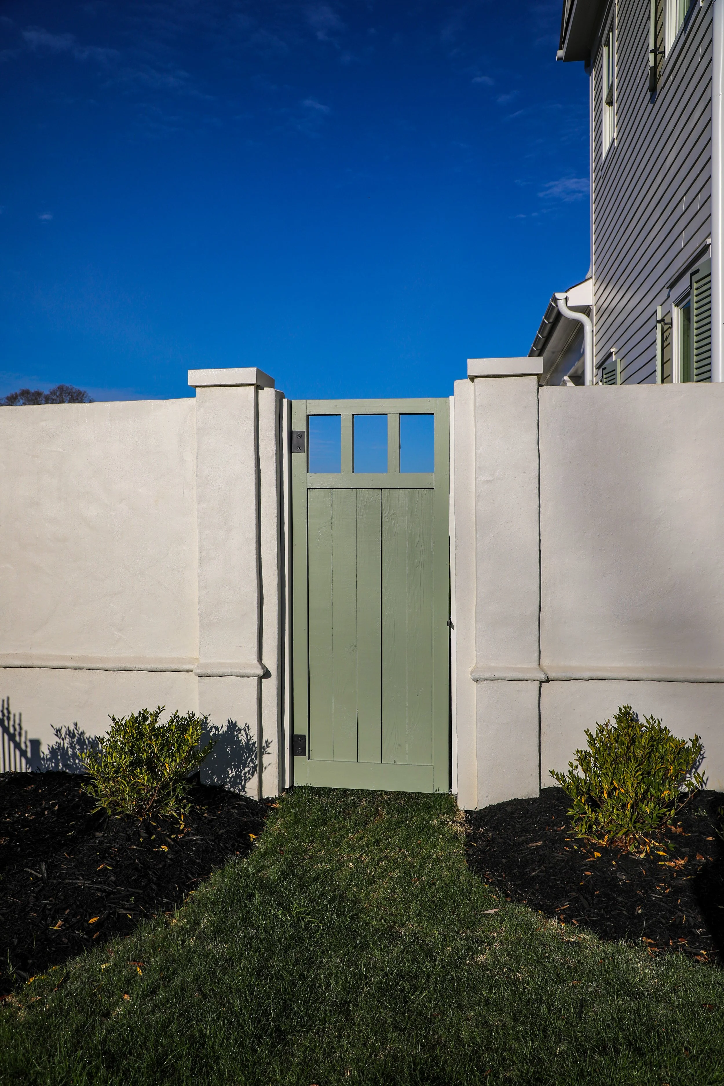 A white stucco fence with a green wooden gate, small shrubs on either side, and a clear blue sky in the background.