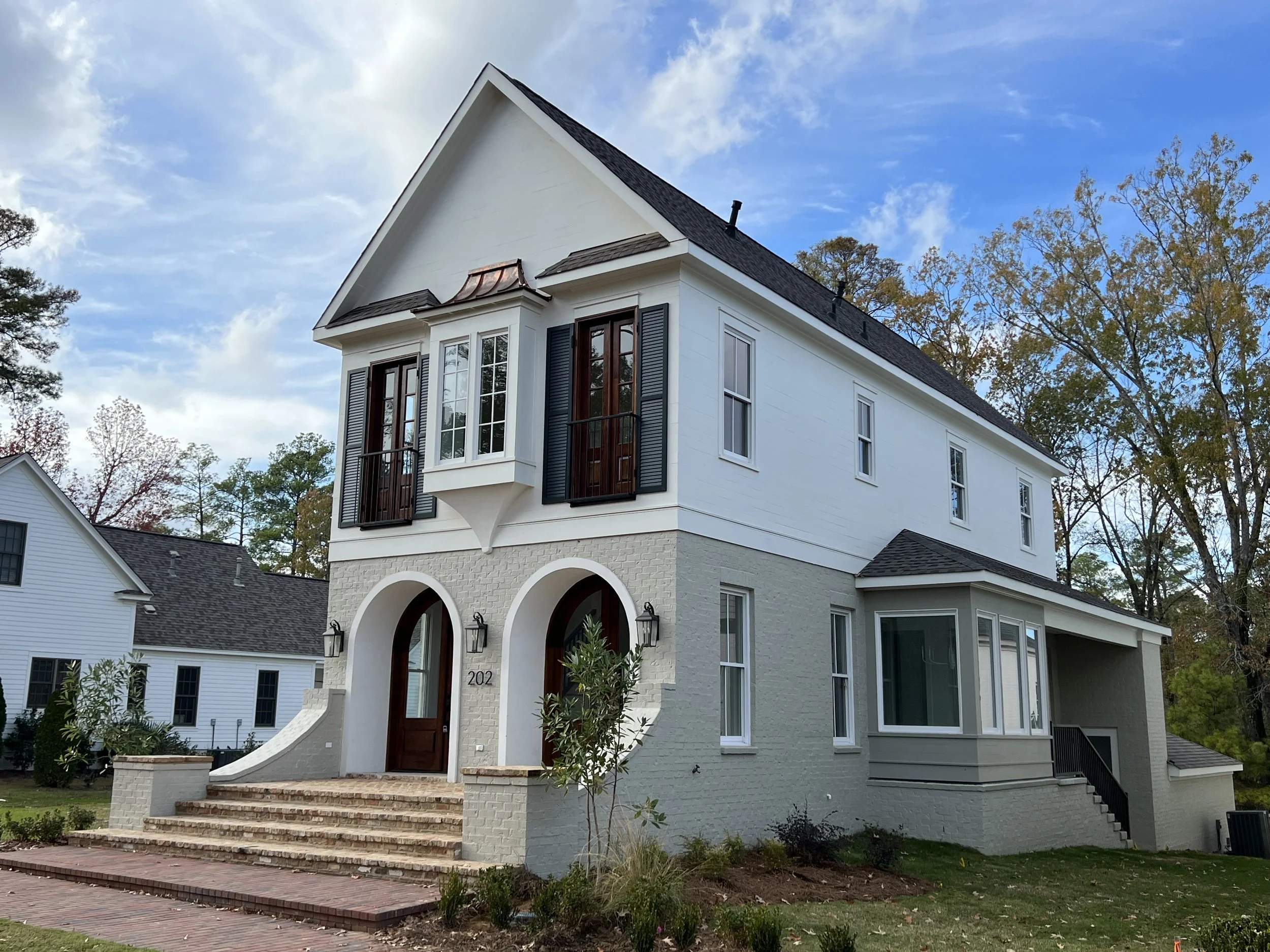 Two-story modern house with brick stairs, white and gray exterior, bay window, and black shutters, surrounded by trees.
