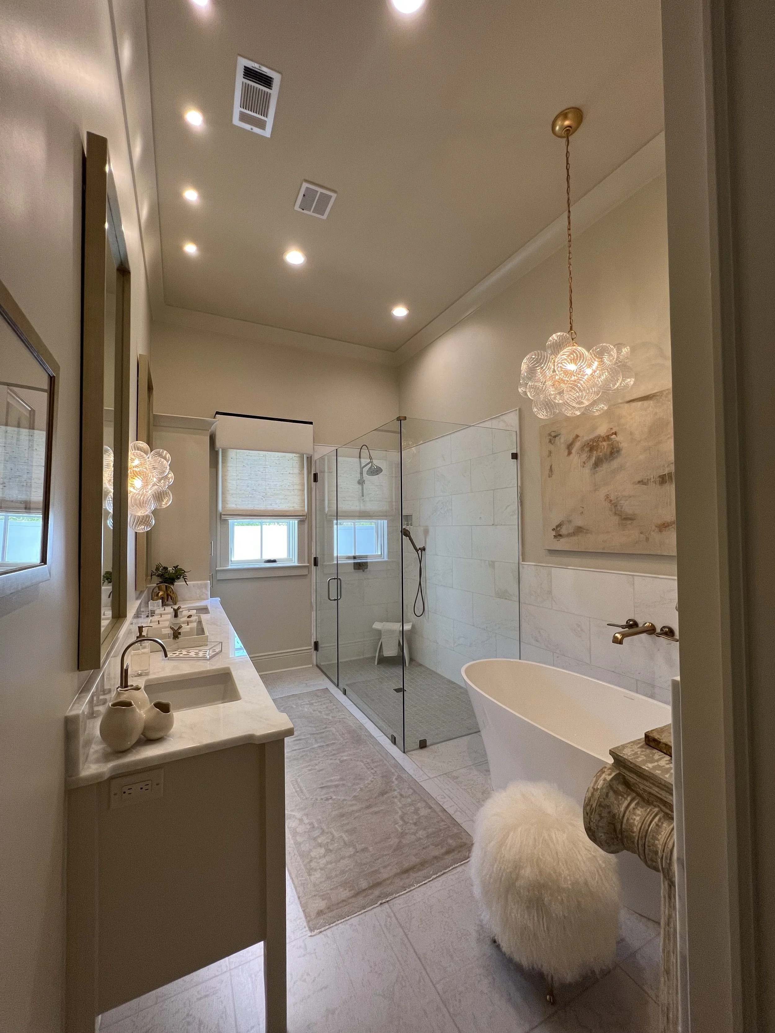 Modern bathroom with dual sinks, a glass-enclosed walk-in shower, a free-standing bathtub, and a fluffy stool. Decor includes framed art, chandeliers, and a beige rug.