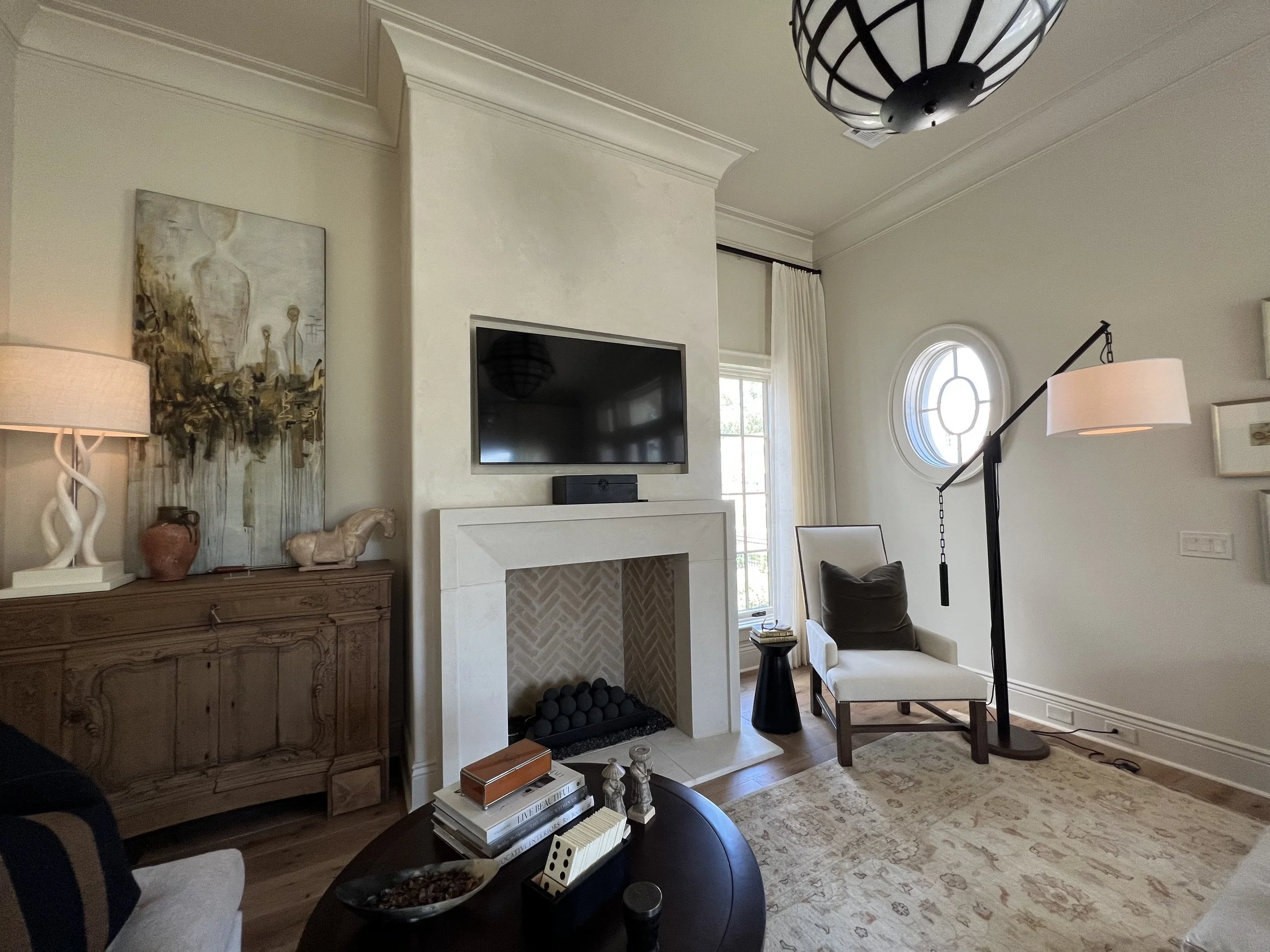 A living room with a mounted flat-screen TV above a fireplace, an art piece on the wall, a wooden console table with decorative items, a chair with a pillow, a tall floor lamp, a window with a curtain, and a beige rug.