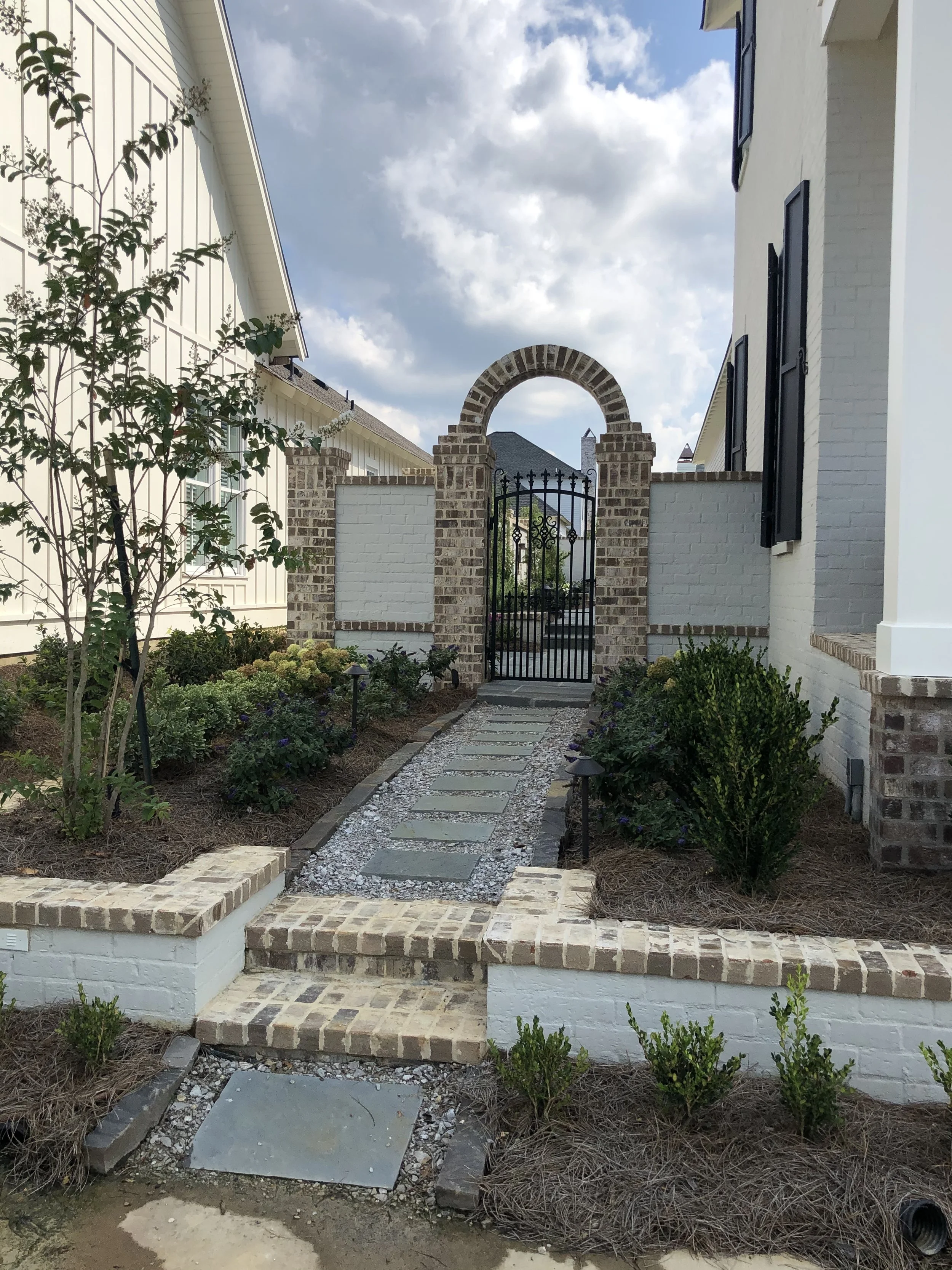A brick pathway leading to a black wrought iron gate surrounded by a brick and white wall, with a garden on either side and a cloudy sky overhead.
