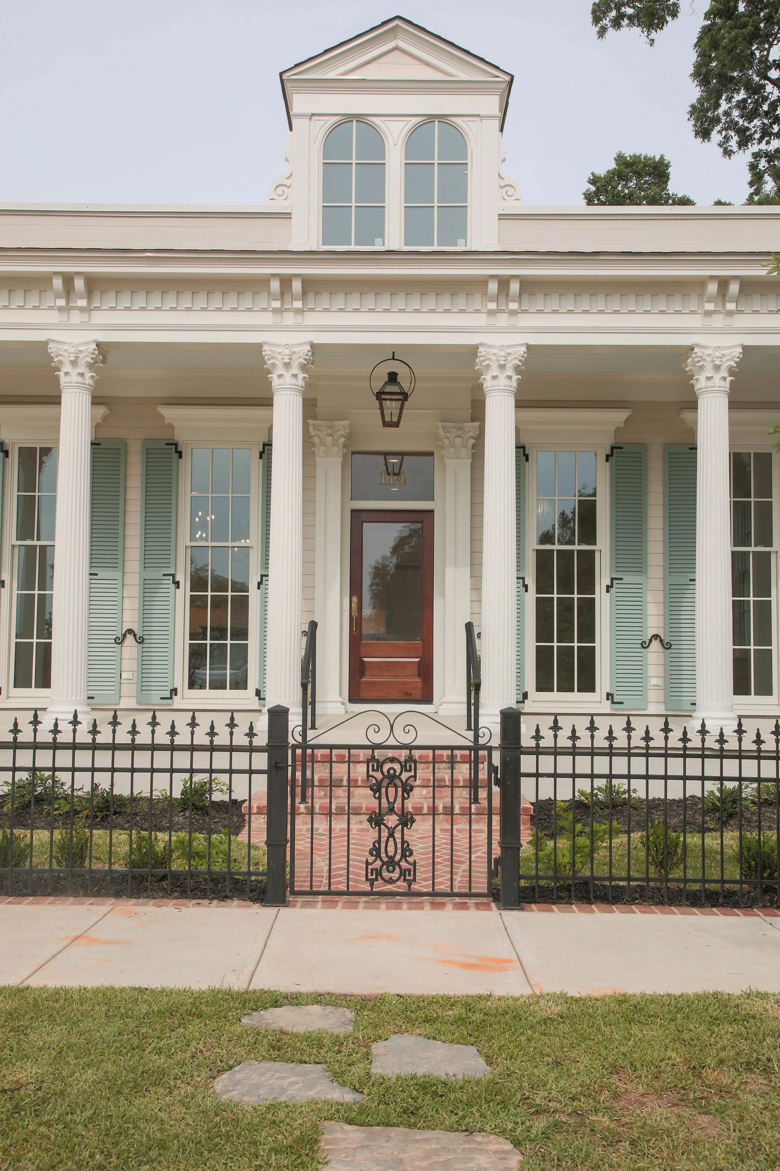 The front exterior of a white, two-story house with tall columns, a small front yard, and a pathway leading to the front door. The house has large windows with blue shutters and decorative trim, and a brick staircase with a wrought iron gate at the e