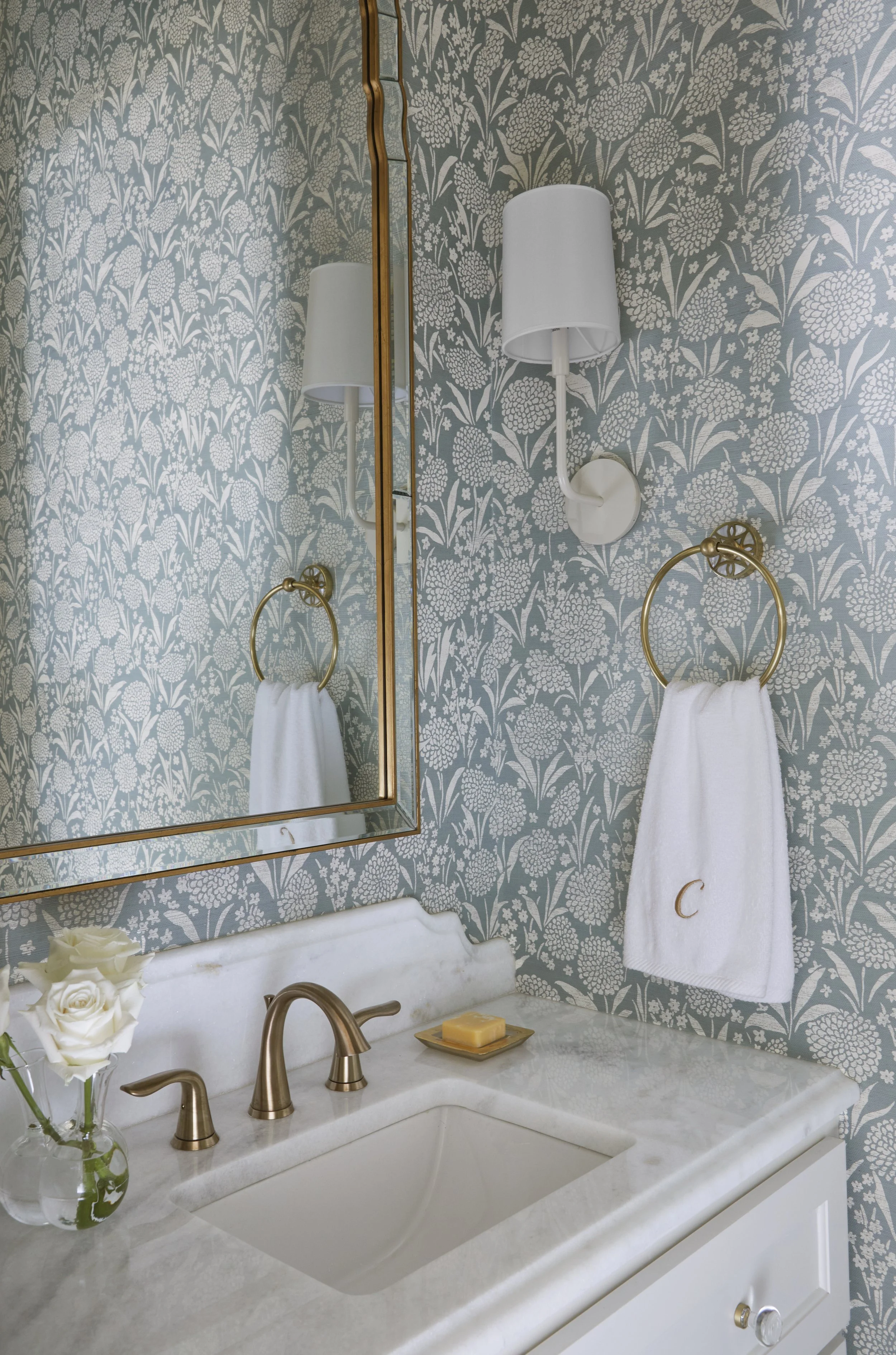 Bathroom with floral wallpaper, a marble countertop, a sink with a bronze faucet, a large mirror, a wall-mounted white lamp, a white towel with a gold letter 'C', a soap dish with soap, and a small vase with white roses.