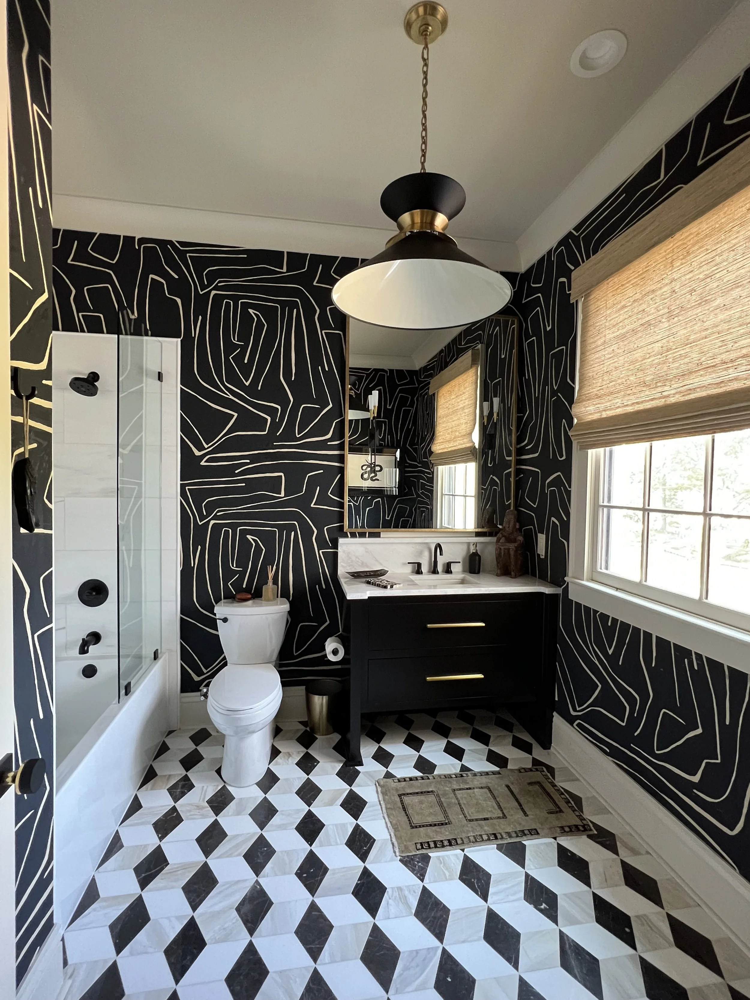 Bathroom with black and white geometric patterned floor, black vanity with gold handles, large mirror, toilet, and walk-in shower with glass door. Black wallpaper with abstract white lines, windows with beige blinds, and a ceiling light fixture.