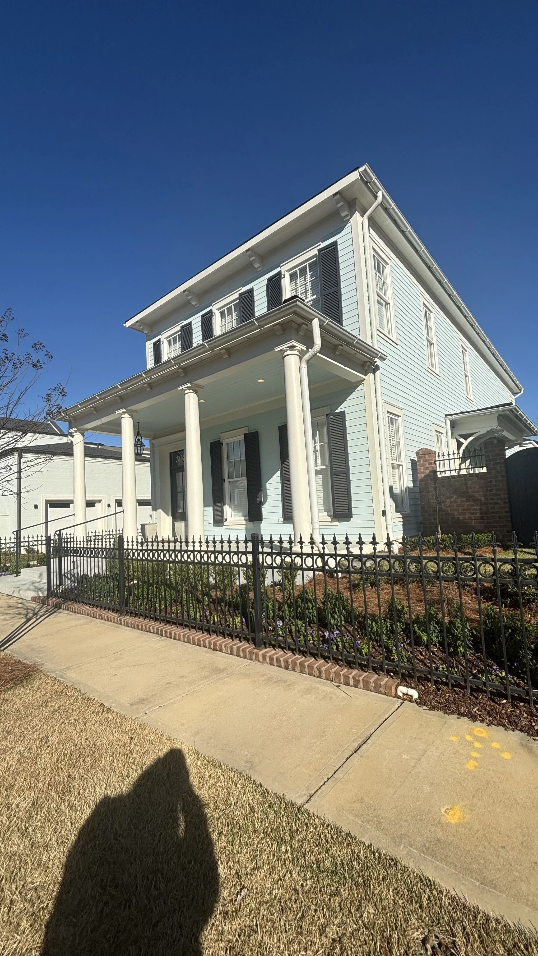 A three-story light blue house with white columns and black shutters, surrounded by a black wrought iron fence, with a sidewalk and grass in the foreground.