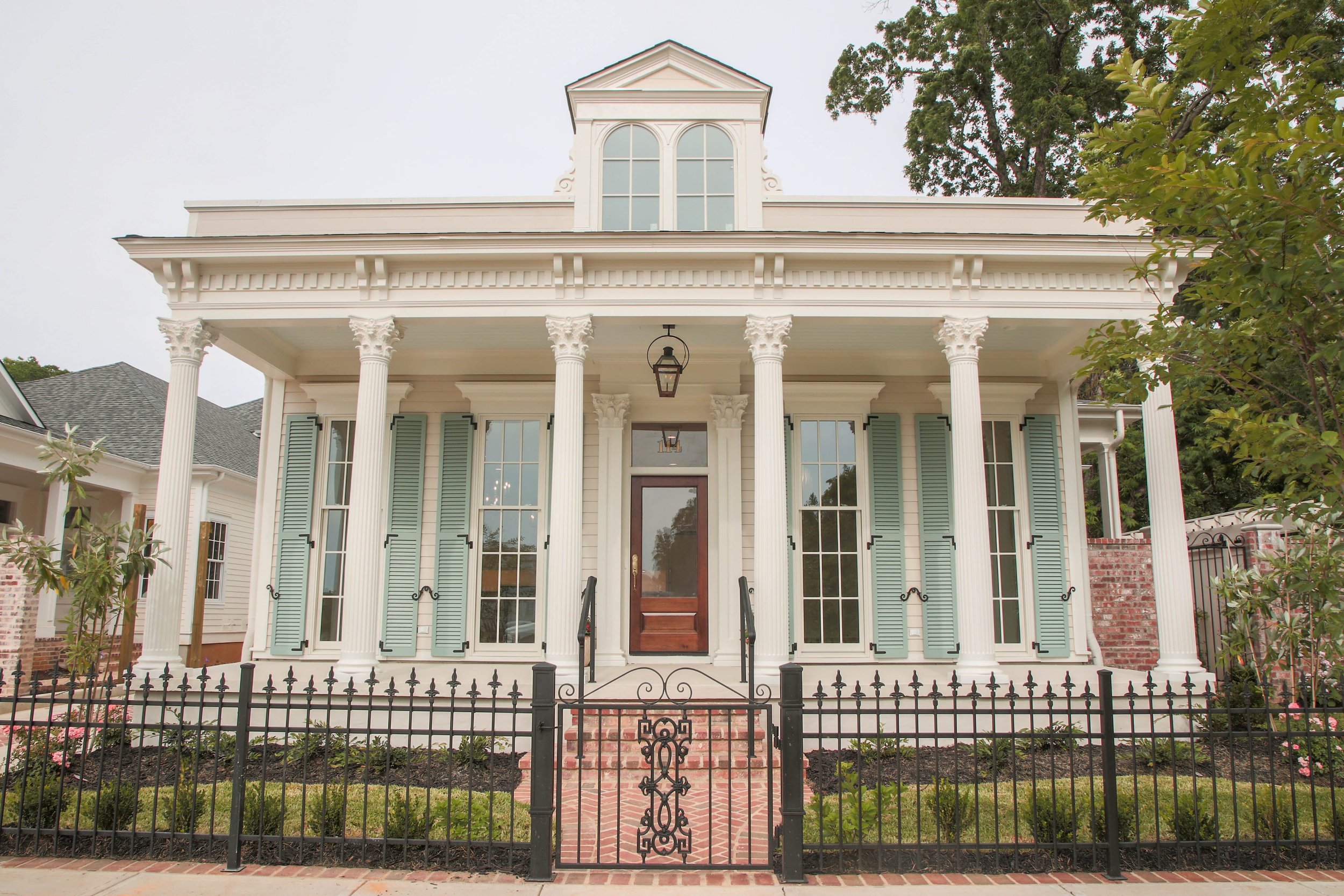 Front view of a white two-story house with green shutters, large columns, and a brick staircase leading to the front door, enclosed by a black iron fence.