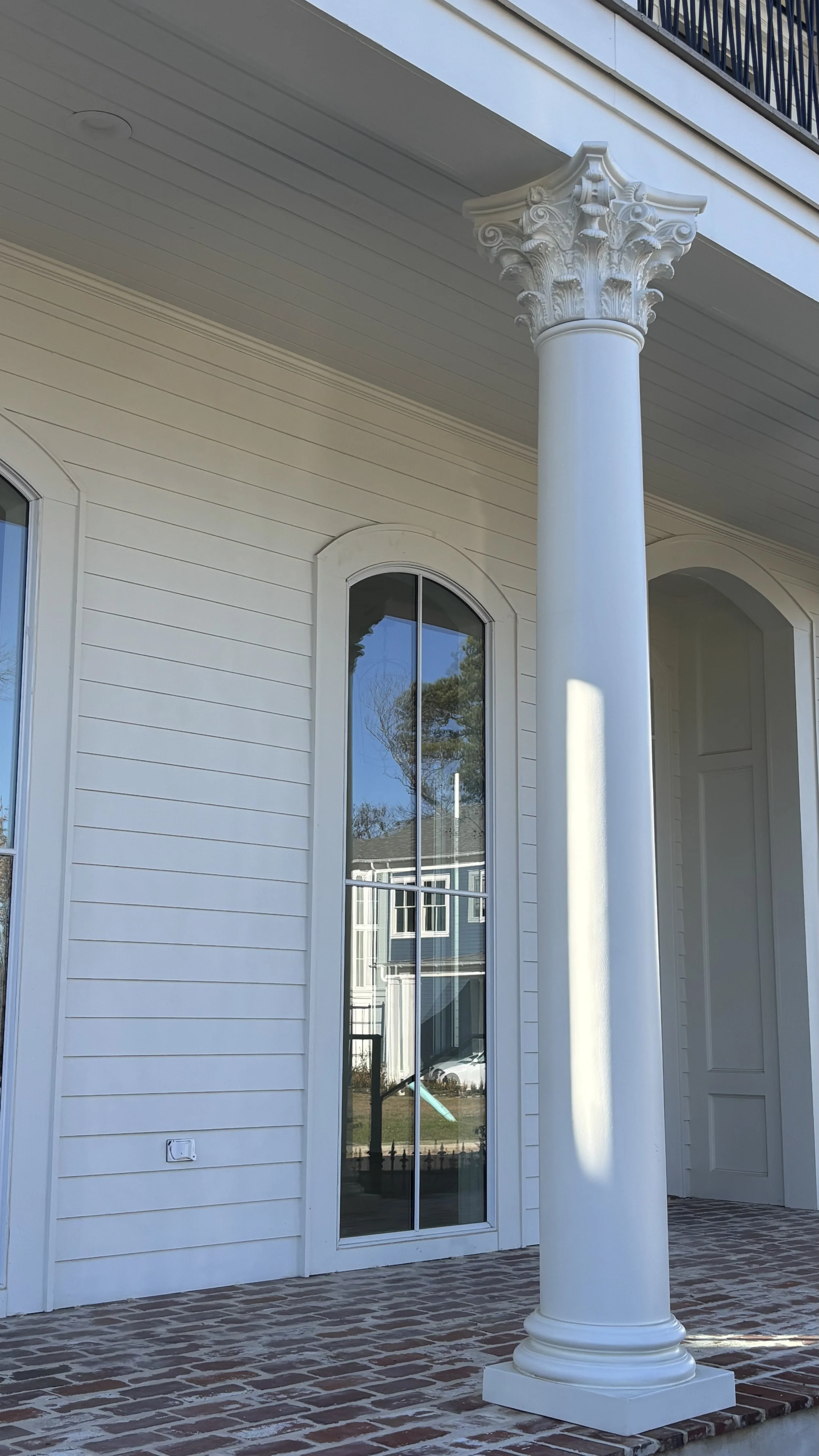 Close-up of white house exterior with large arched window, decorative white column with ornate capital, brick porch, and siding.