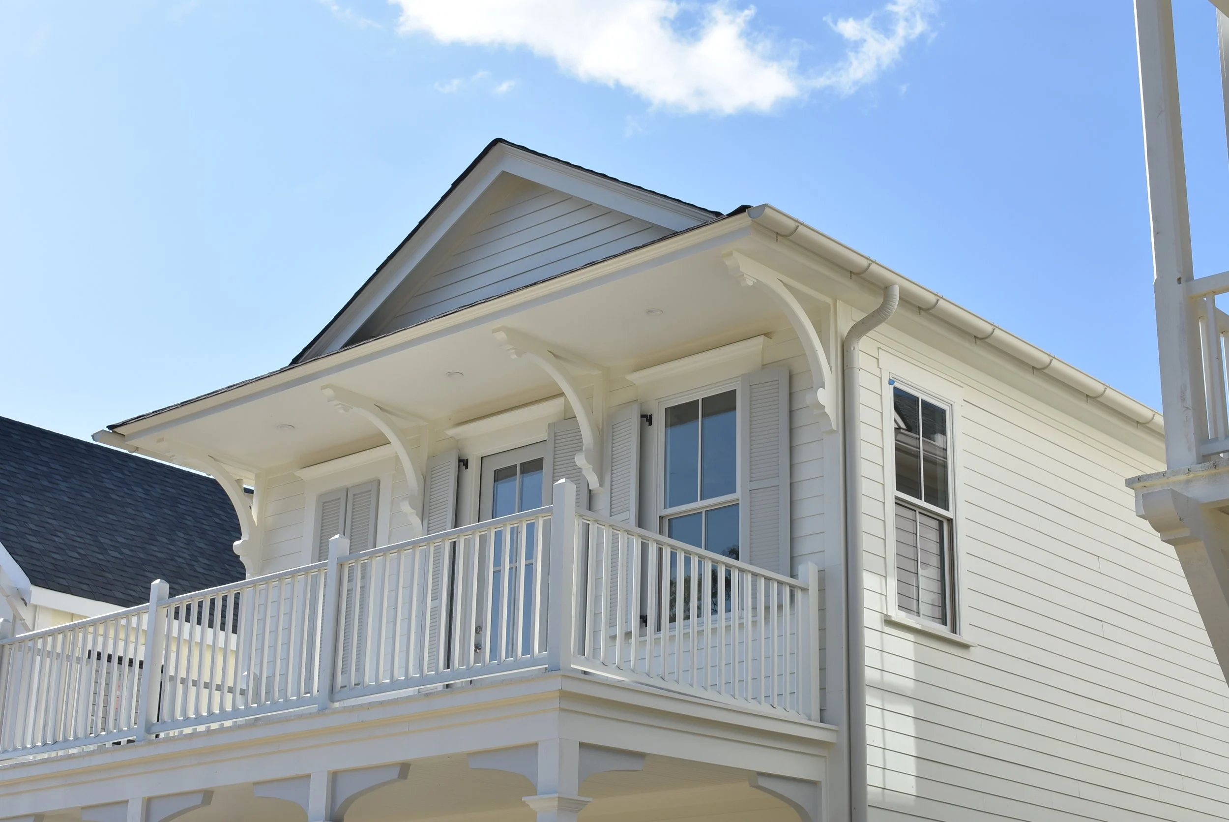 A white two-story house with a small balcony, window shutters, and a gabled roof against a blue sky with some clouds.