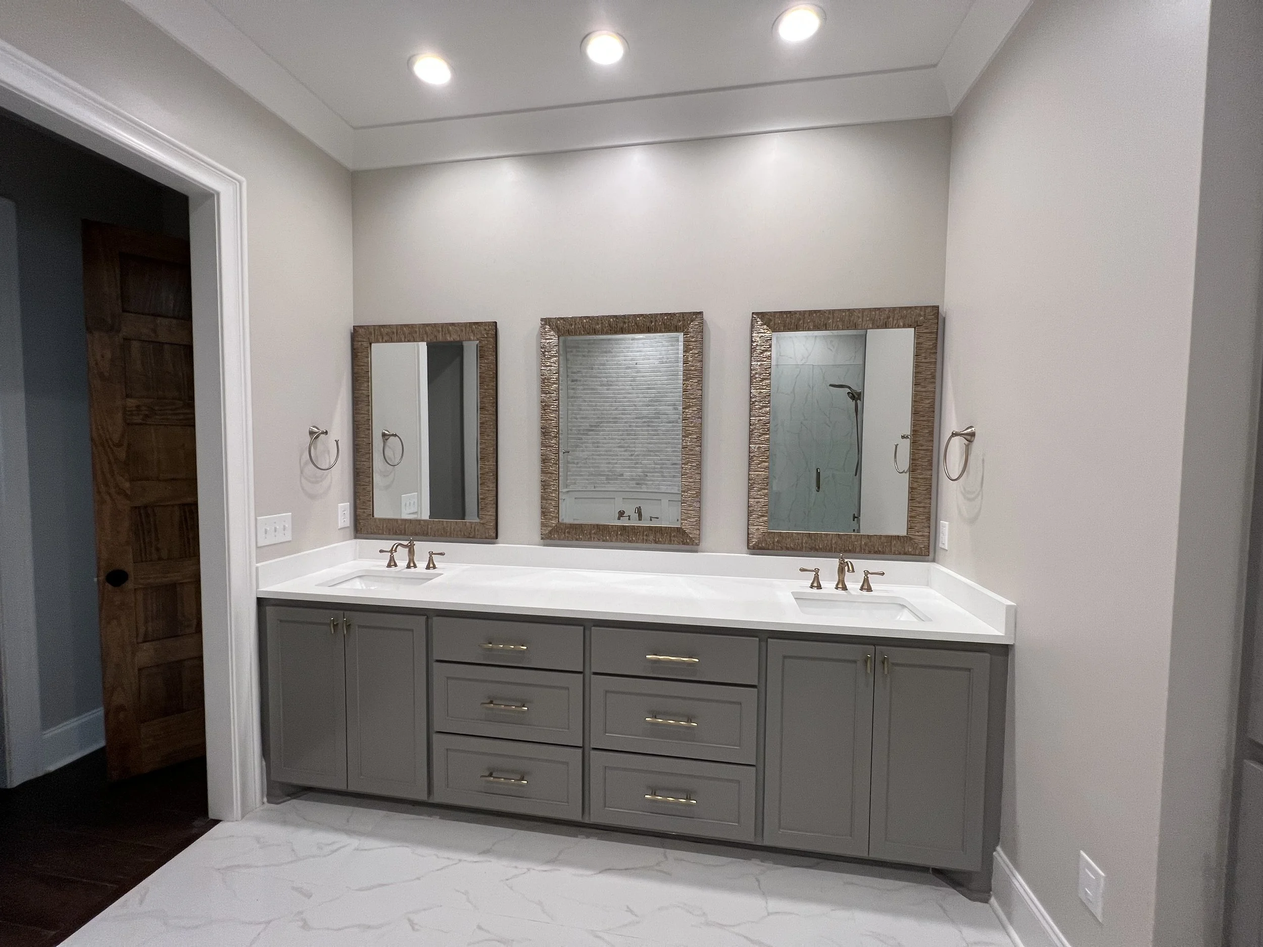 A double bathroom vanity with gray cabinets, white countertop, and two sinks with brass fixtures. Above, there are three framed mirrors. The bathroom has beige walls, recessed ceiling lights, and a white marble-like floor.