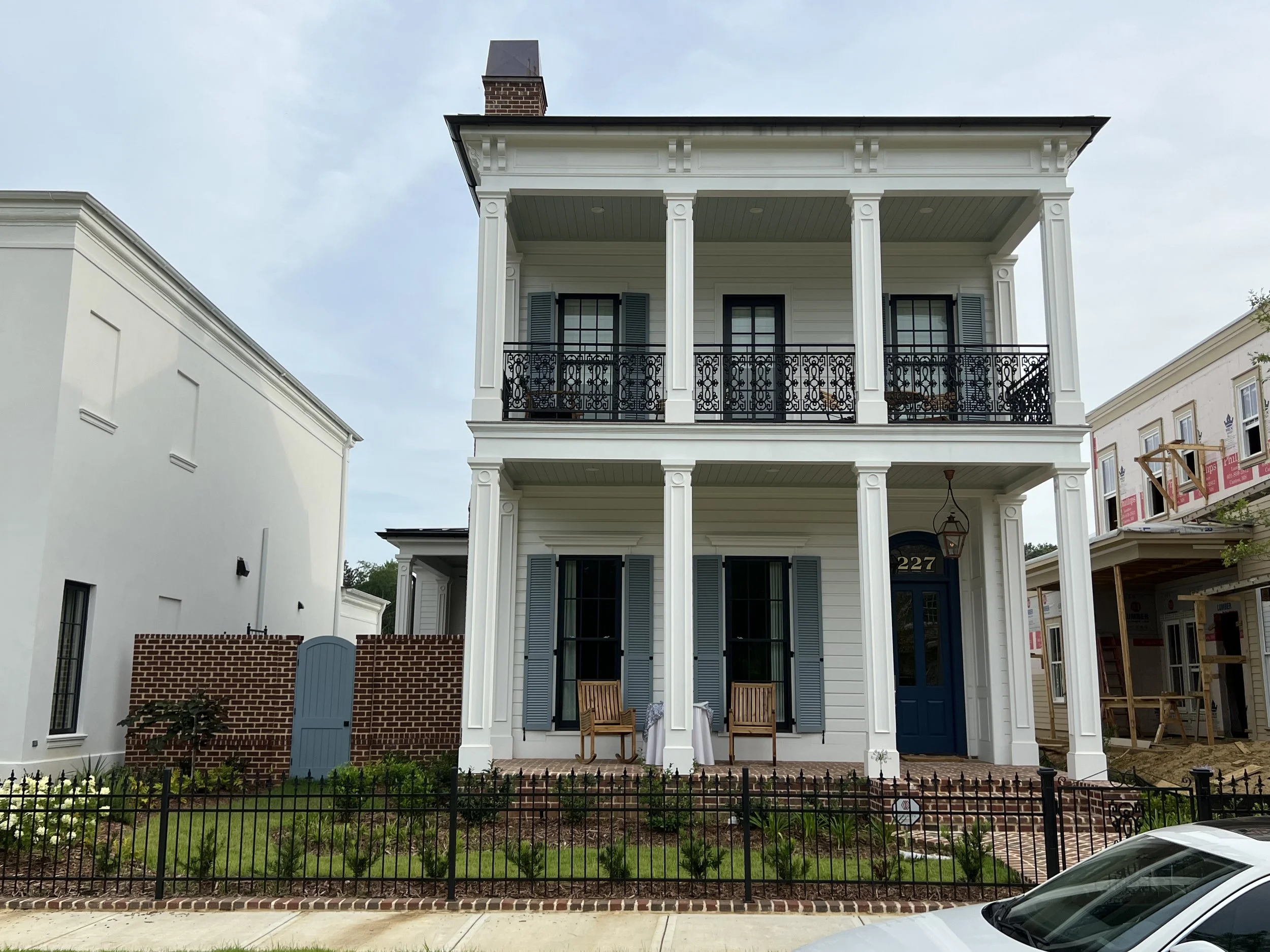 Two-story white house with columns, black balconies, blue shutters, and a blue door, with a small front yard and a black fence. There is a table and two chairs on the front porch.