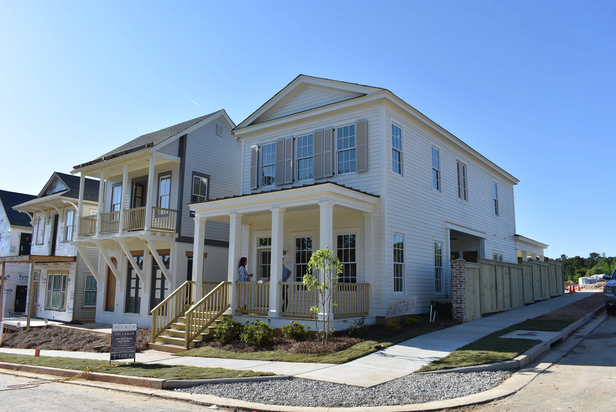 Newly constructed white multi-story house with front porch, stairs, and windows, under a clear blue sky, in a residential neighborhood with neighboring houses and construction signs.