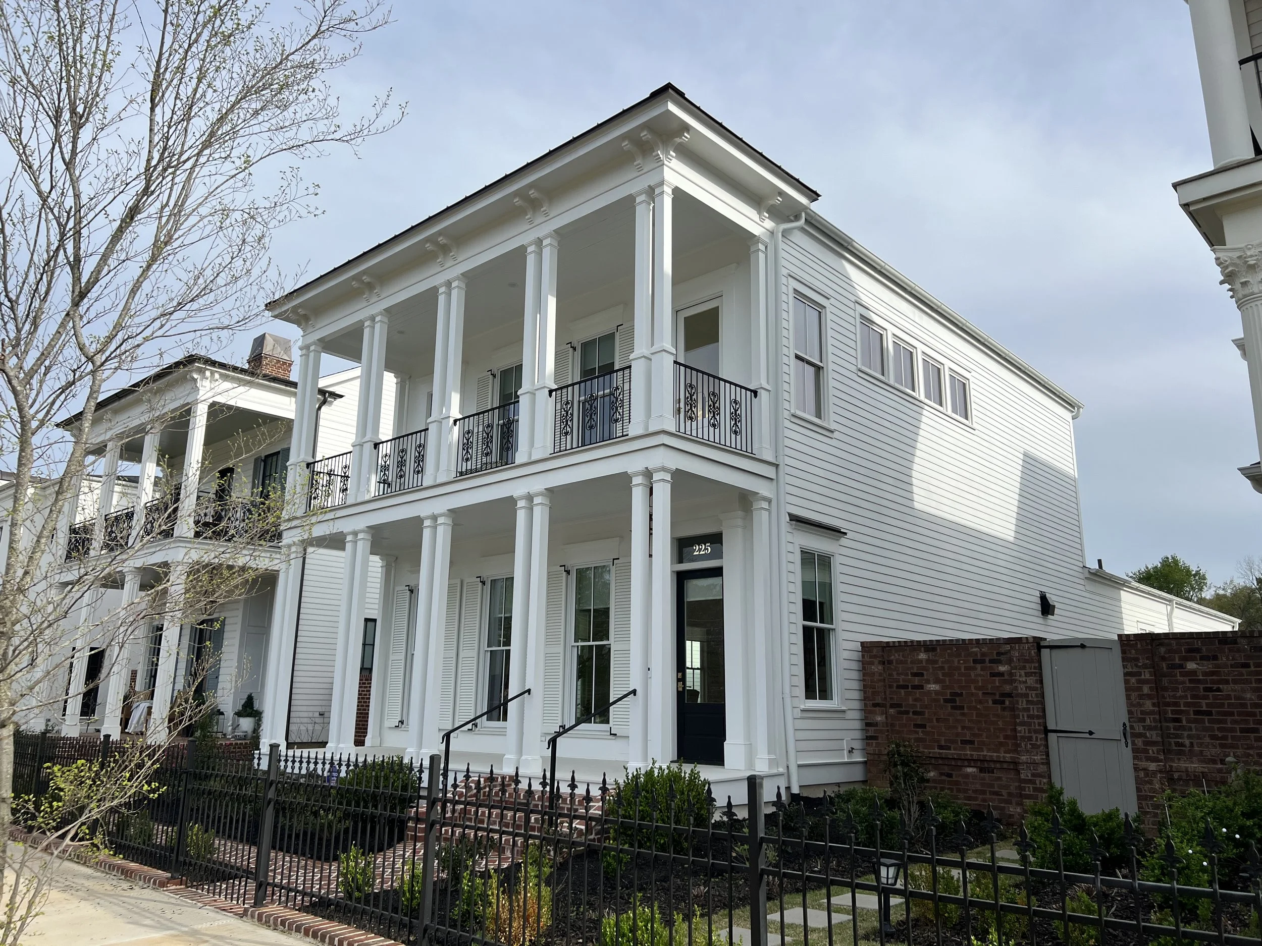White, three-story residential building with black balconies and columns, black front door, and surrounded by a black fence, with a brick wall on one side.
