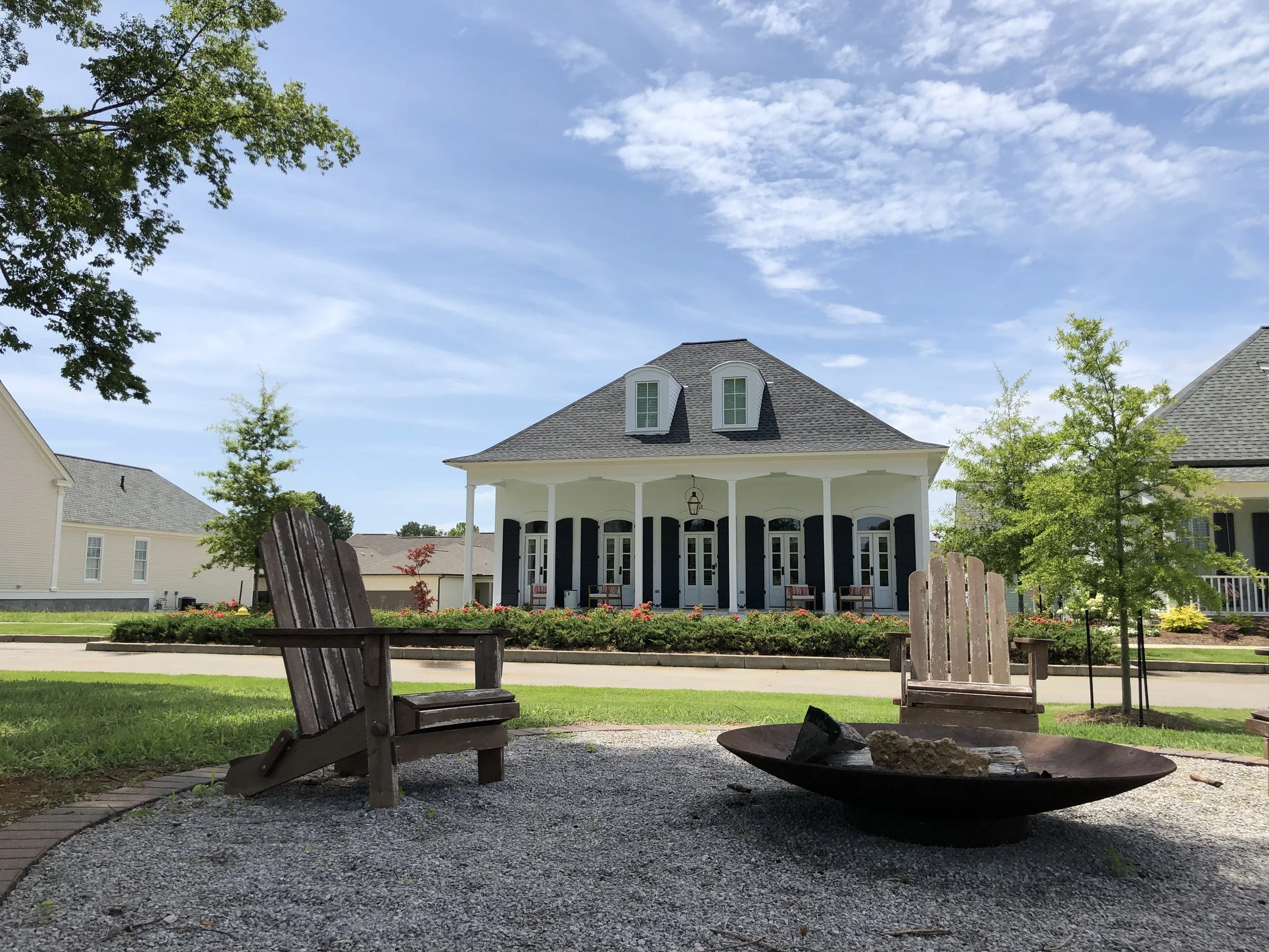 Front view of a large white house with black shutters and a porte-cochère, surrounded by trees and a landscaped yard, under a blue sky with some clouds. In the foreground, there are two wooden chairs and a fire bowl with rocks.