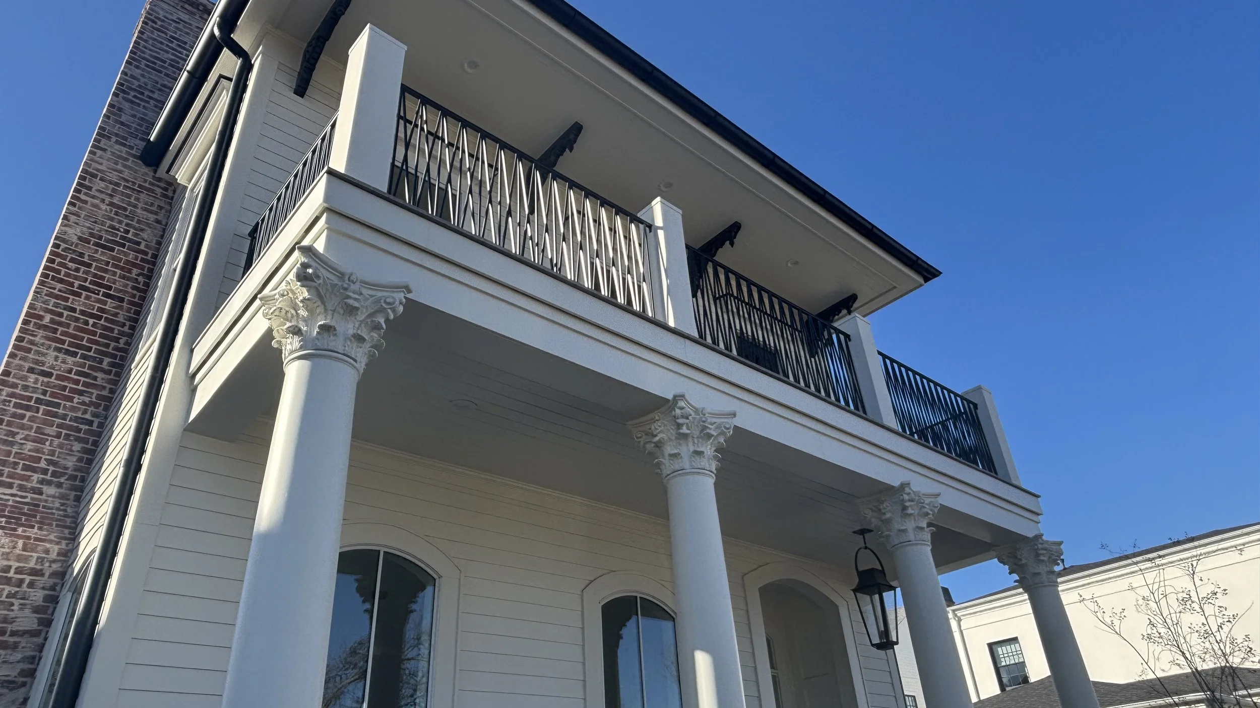View of a modern multi-story house with classical columns supporting the front porch and balcony. The house has white siding, black railings, and large windows, with a clear blue sky in the background.