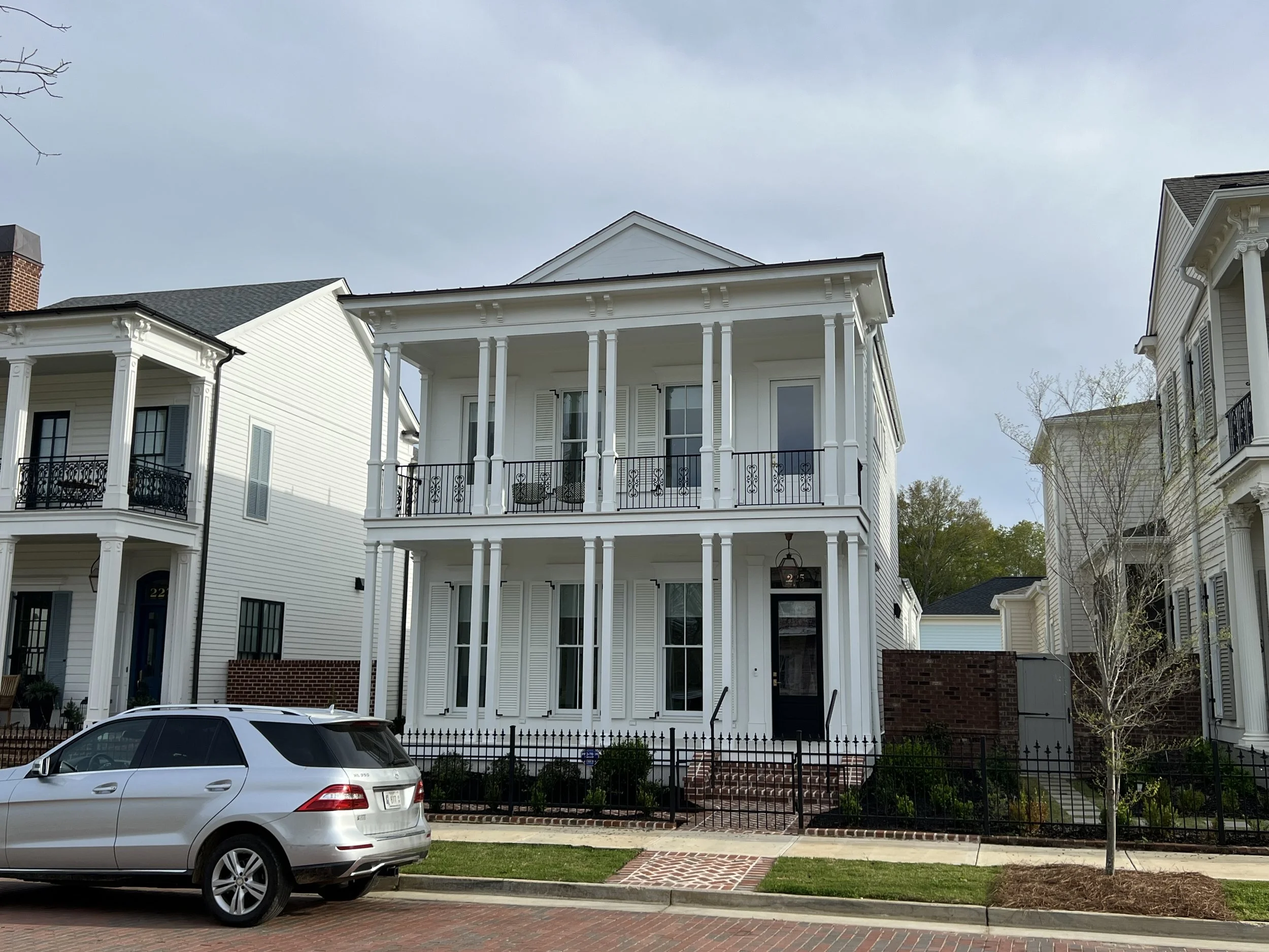 A three-story white house with a covered front porch, black railings, large windows with shutters, and brick stairs leading to the front door. There are neighboring houses on either side, a black fence, and a silver car parked in front.