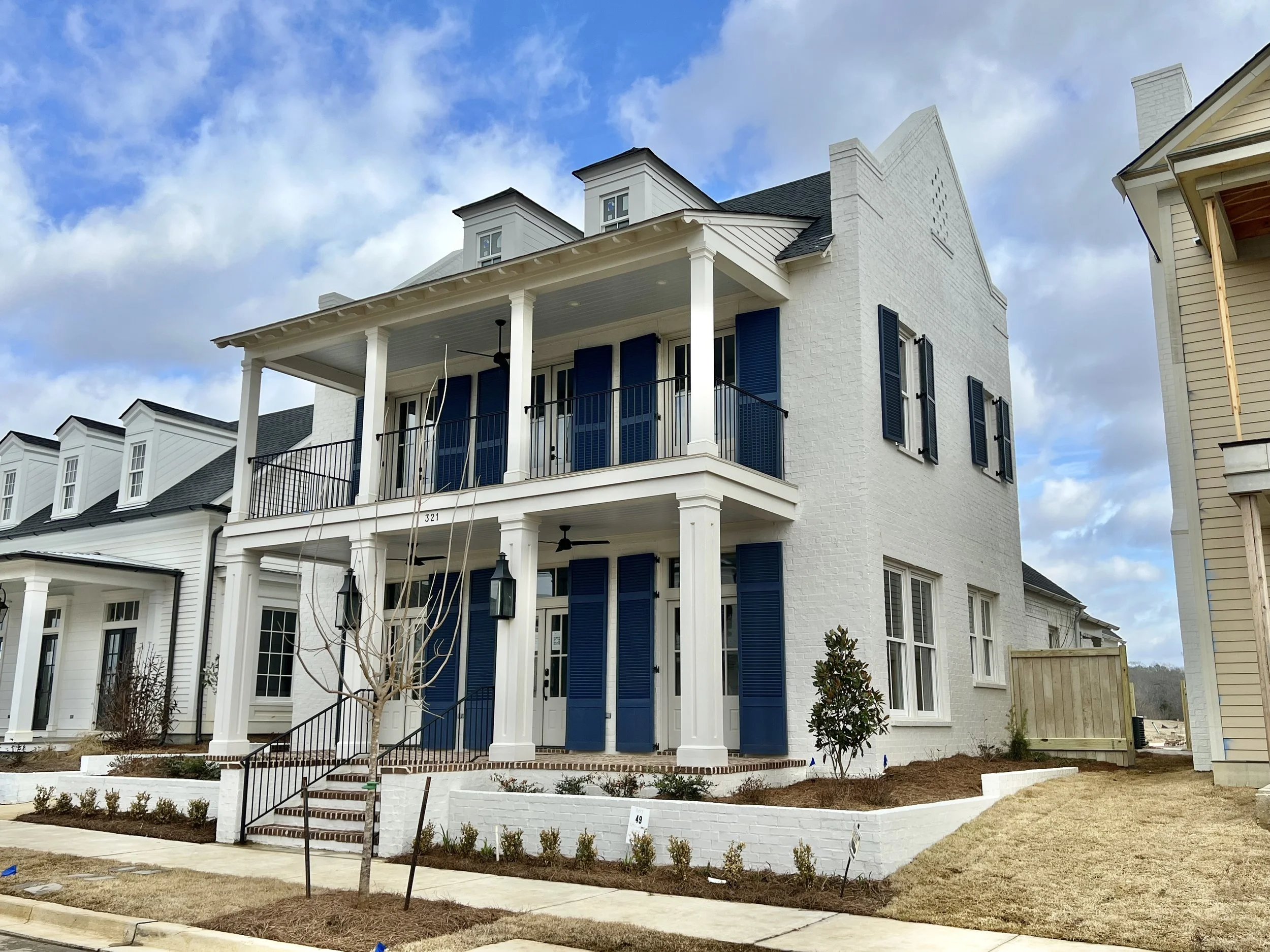 A modern white multi-story house with blue shutters, a front porch with black railings, and a landscaped yard with small plants and a young tree.