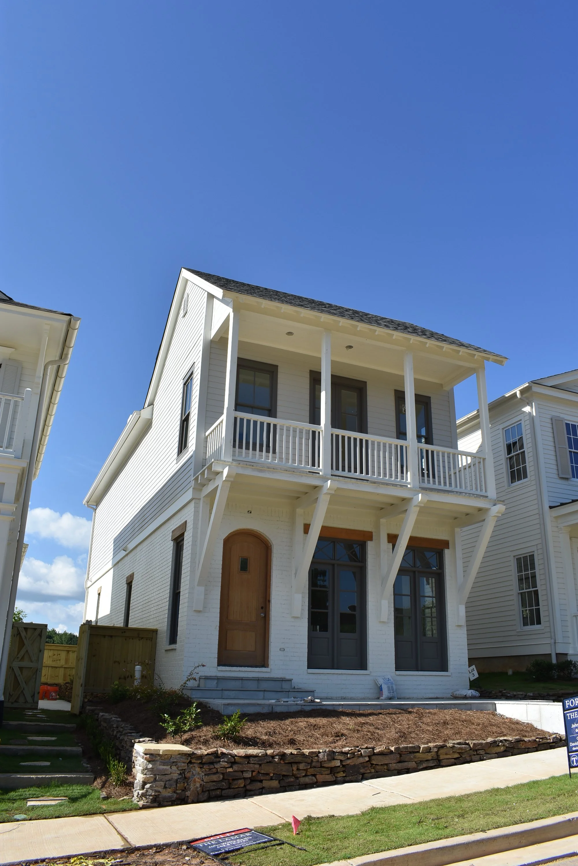 Newly constructed white house with a wooden front door, large windows, and a second-story balcony under a clear blue sky.