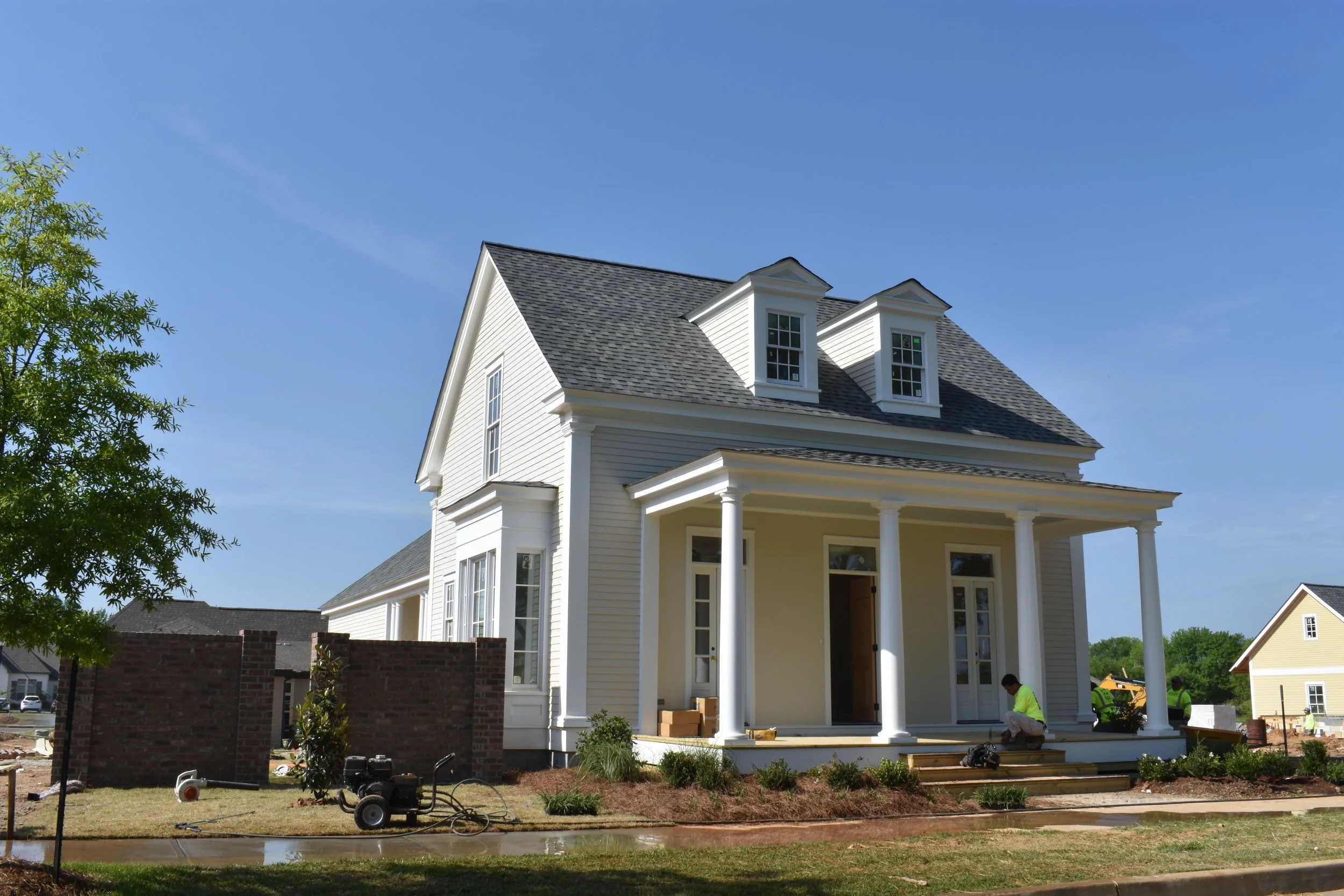A new white two-story house under construction with a porch supported by columns, workers in neon vests working on the front steps, a brick wall, and construction equipment in the yard on a clear day.