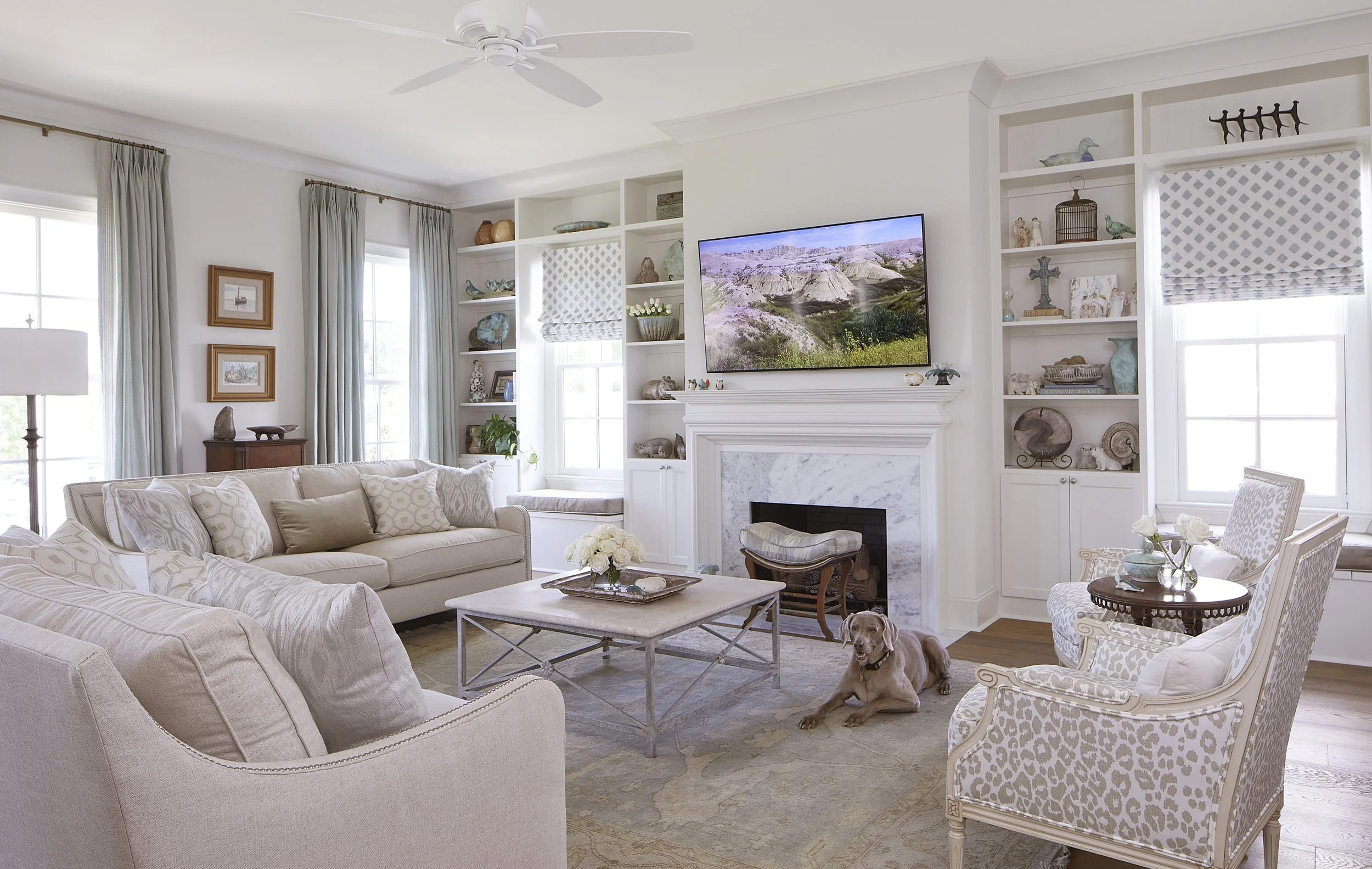 Bright living room with white walls, overlooking shelves with decorative items, a fireplace, and a mounted TV. Features beige and white furniture, a dog lying on the rug, and large windows with curtains.