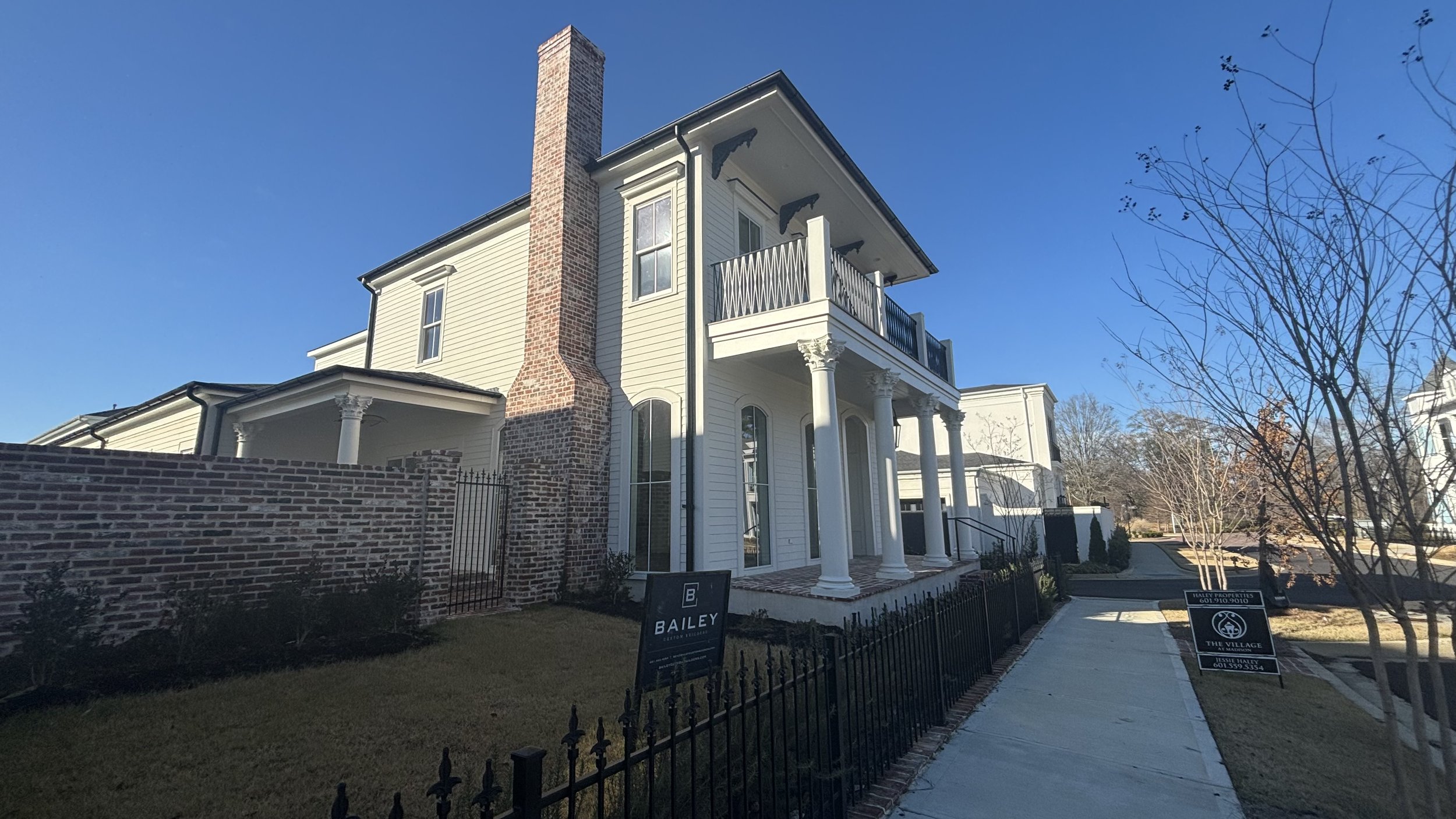 A multi-story house featuring a white exterior, tall brick chimney, and decorative columns on porch, with a sidewalk and some trees in the neighborhood.