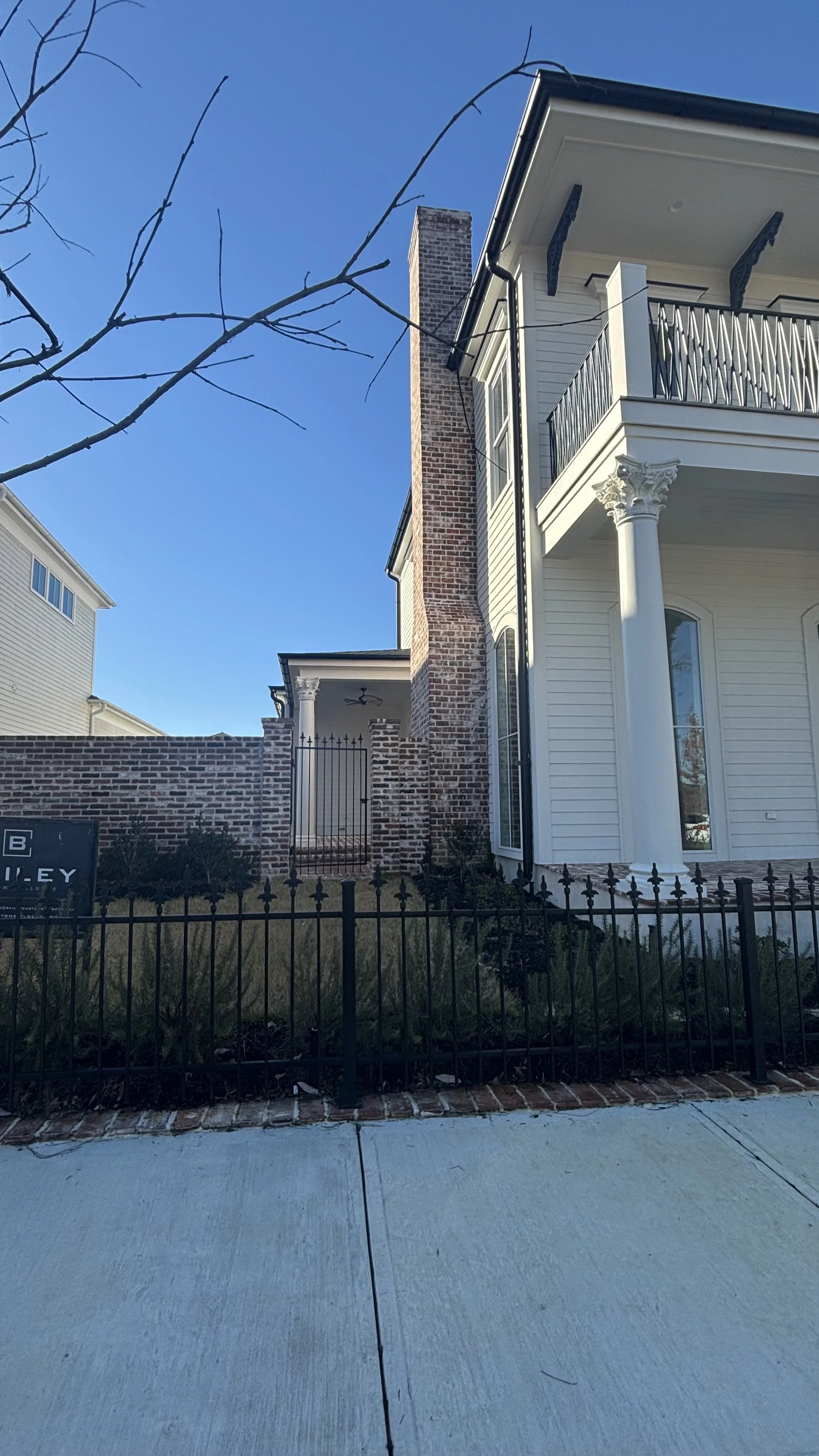 Front view of a modern house with a brick chimney, white siding, columns, and a balcony, surrounded by a black metal fence, with a clear blue sky.