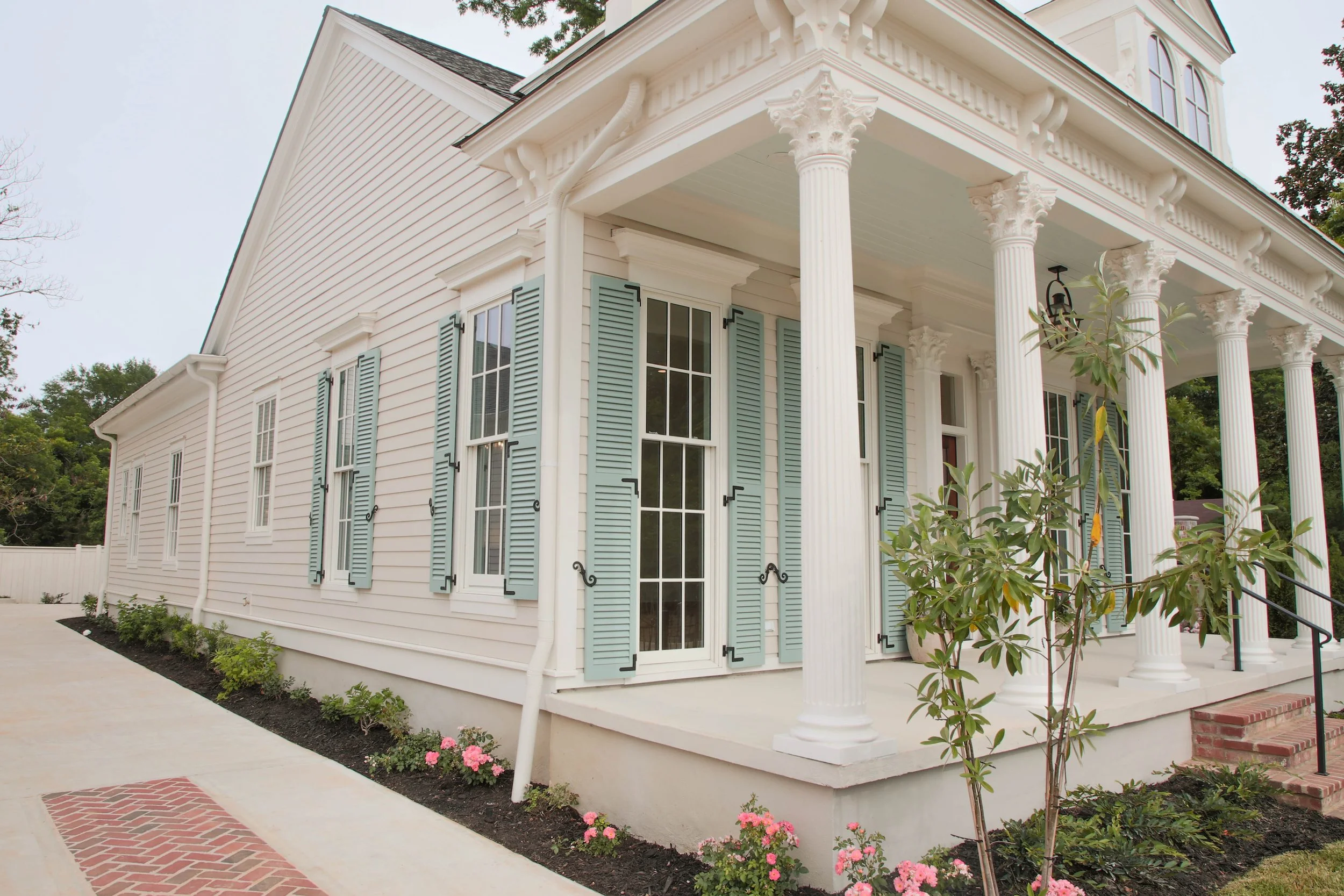 Exterior of a white house with green shutters and classical columns on the porch. There are pink flowers and a small tree in front.