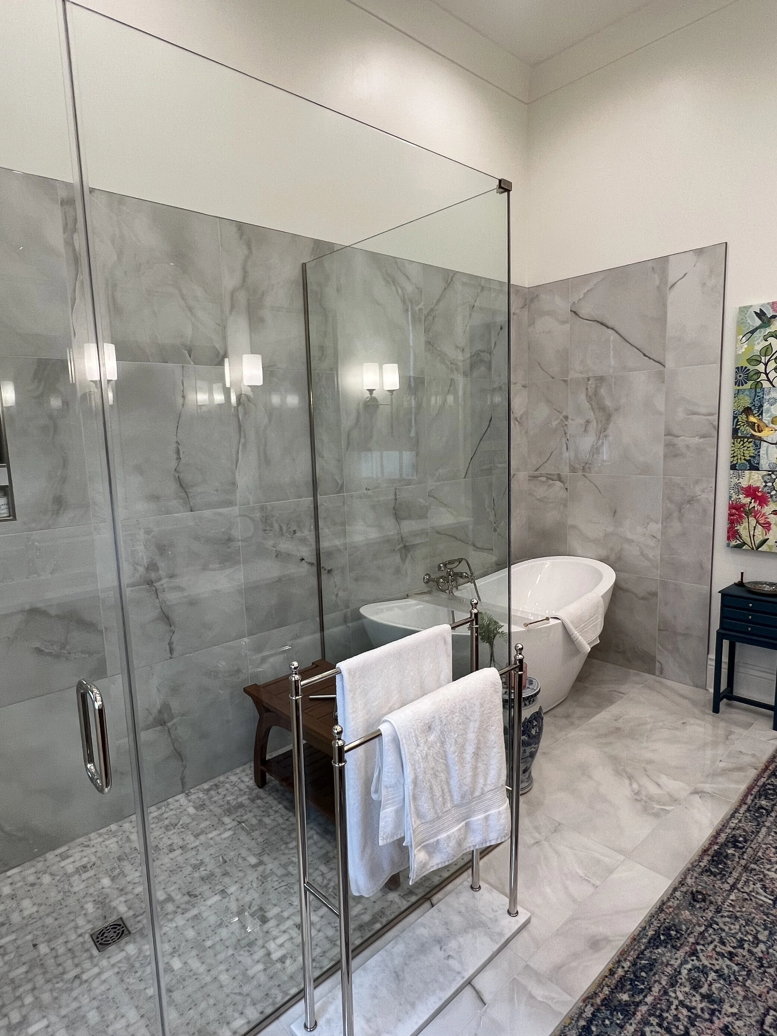 A bathroom with marble walls and floor, a glass enclosed shower, a vintage white bathtub, a towel rack with towels, a wooden chair, and decorative artwork.