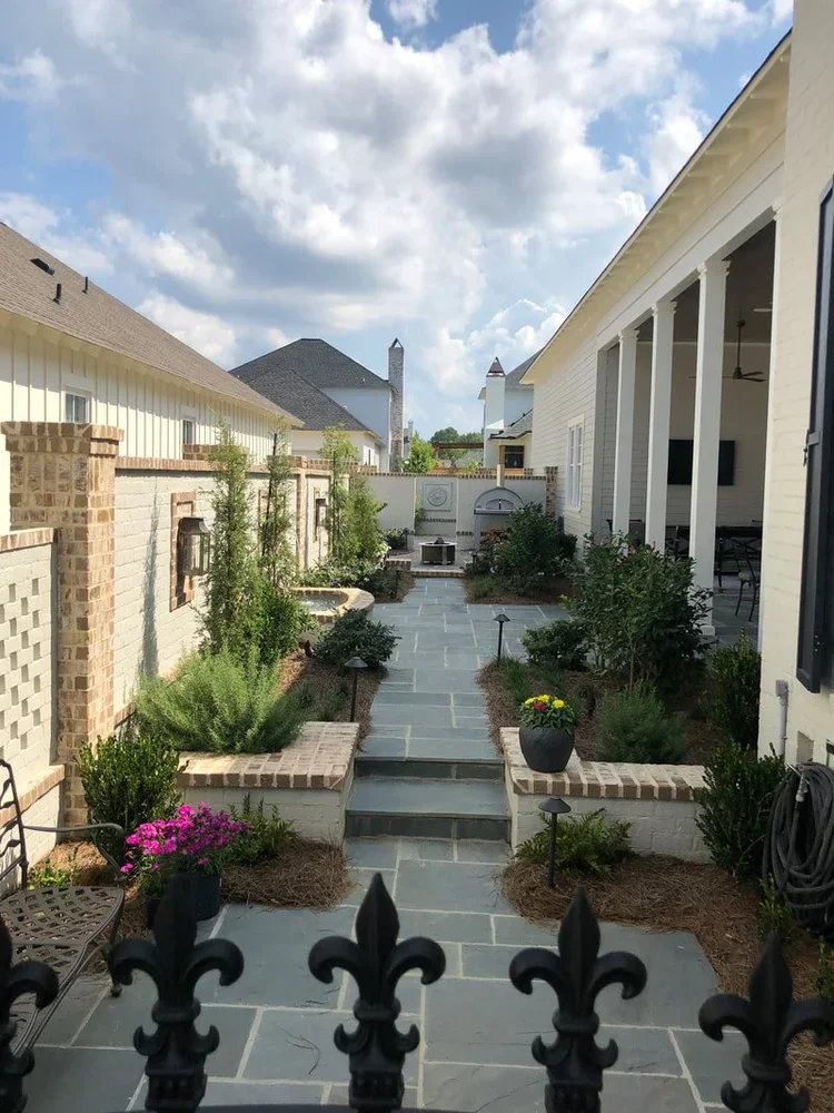 A backyard patio with a stone walkway, lush green plants, bushes, and flowers, surrounded by a brick and white wall, with houses and a partly cloudy sky in the background.