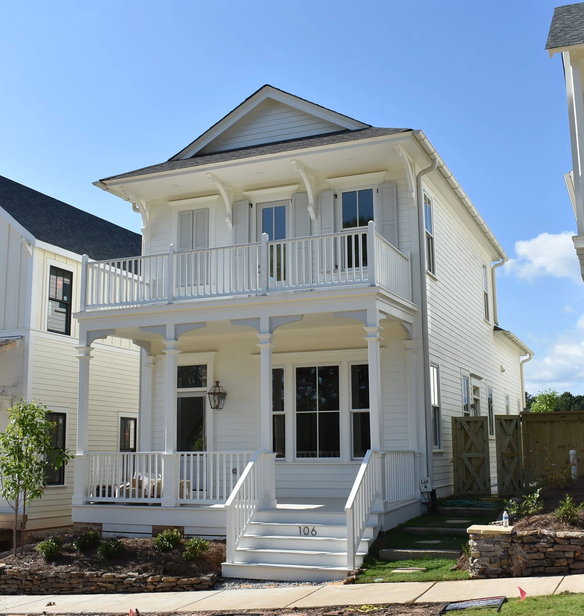White three-story house with a front porch and balcony, surrounded by a landscaped yard with small plants and a young tree, under a clear blue sky.