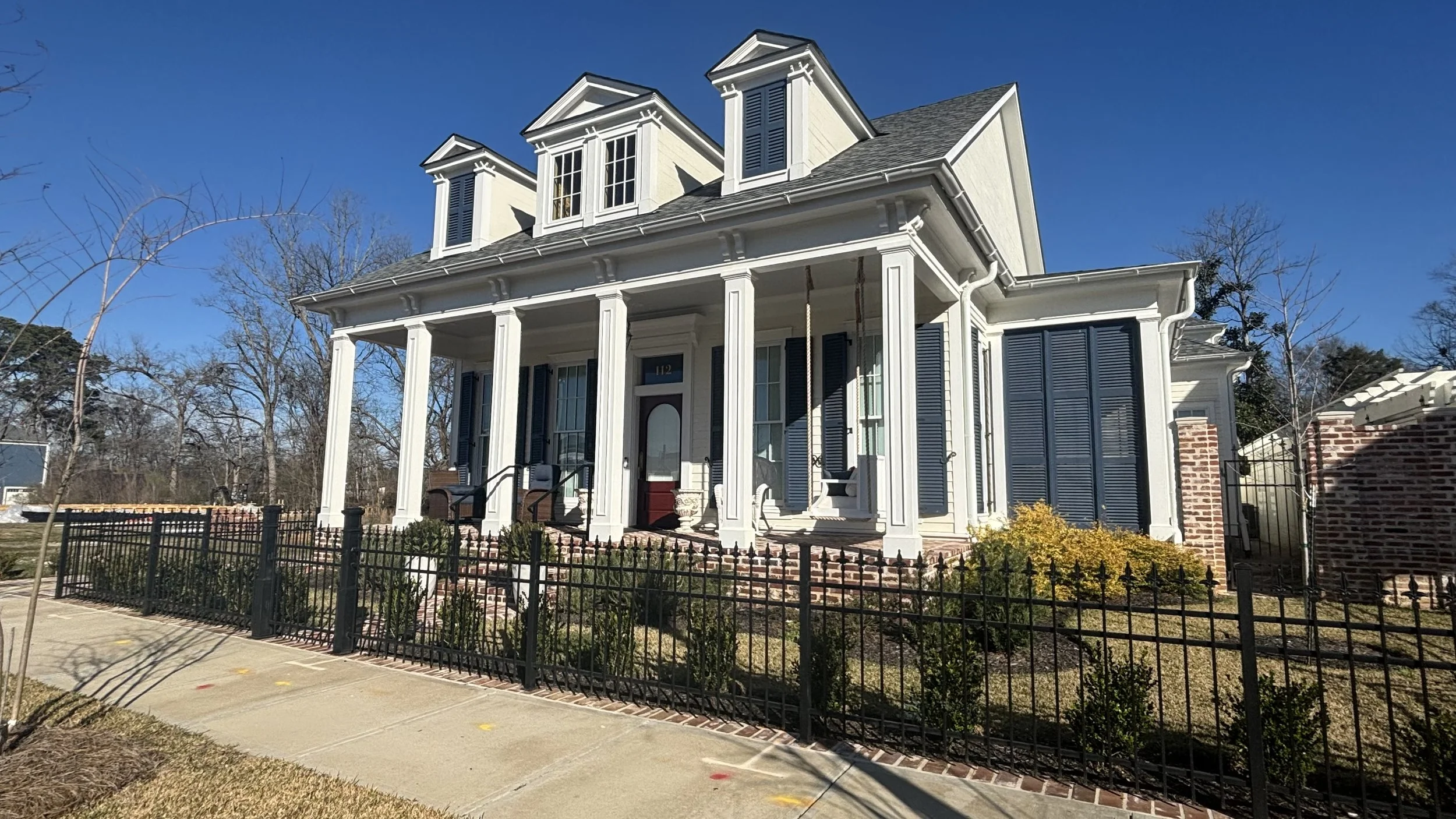 A large white house with three front-facing dormer windows, black shutters, a red front door, and a covered porch with black railings. The house has a brick staircase and is surrounded by a black metal fence. There is a sidewalk in front of the house