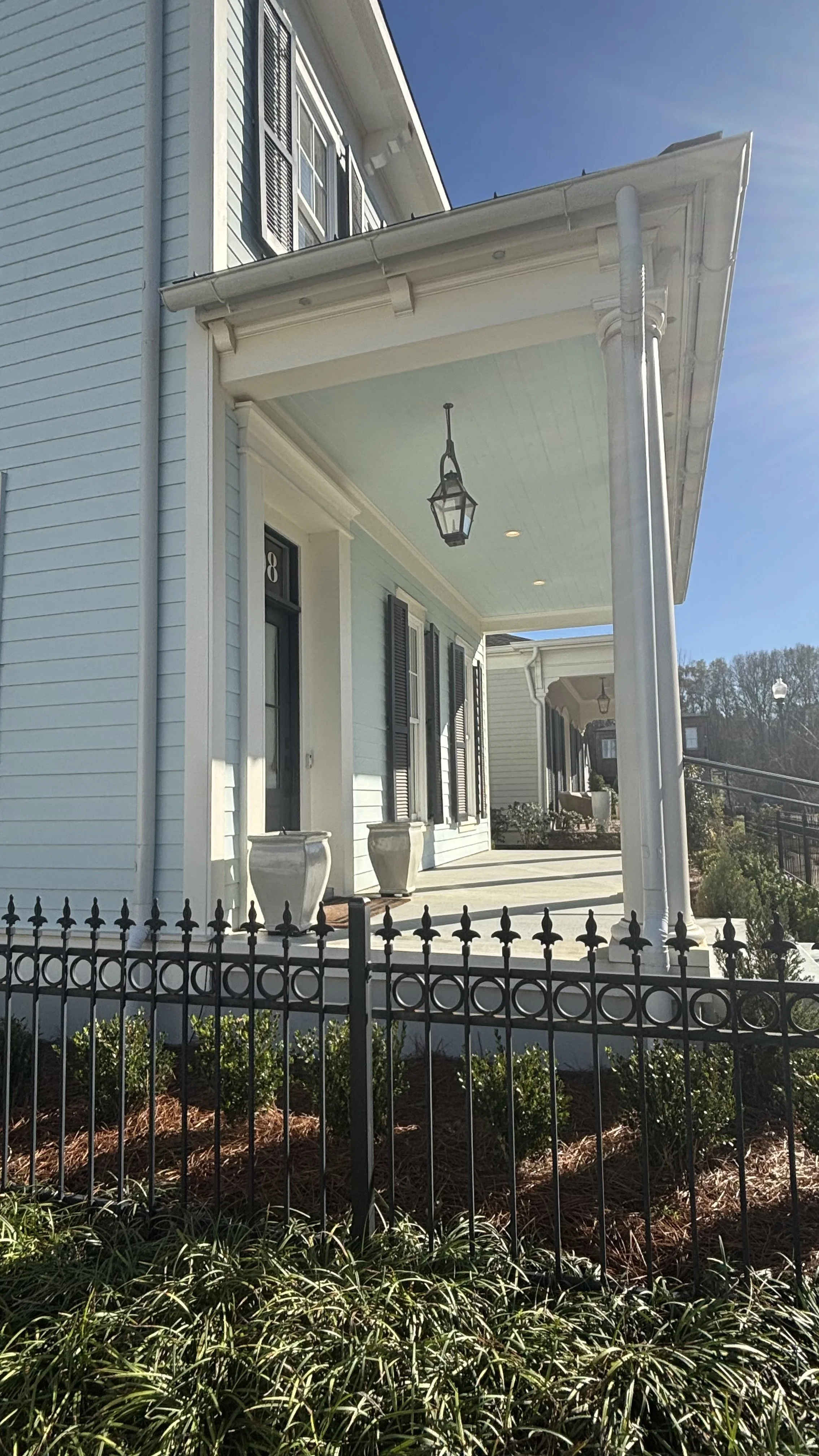 Front porch of a light blue house with white trim, black shutters, two large potted plants, a black fence, and a walkway with plants and mulch in the garden bed.