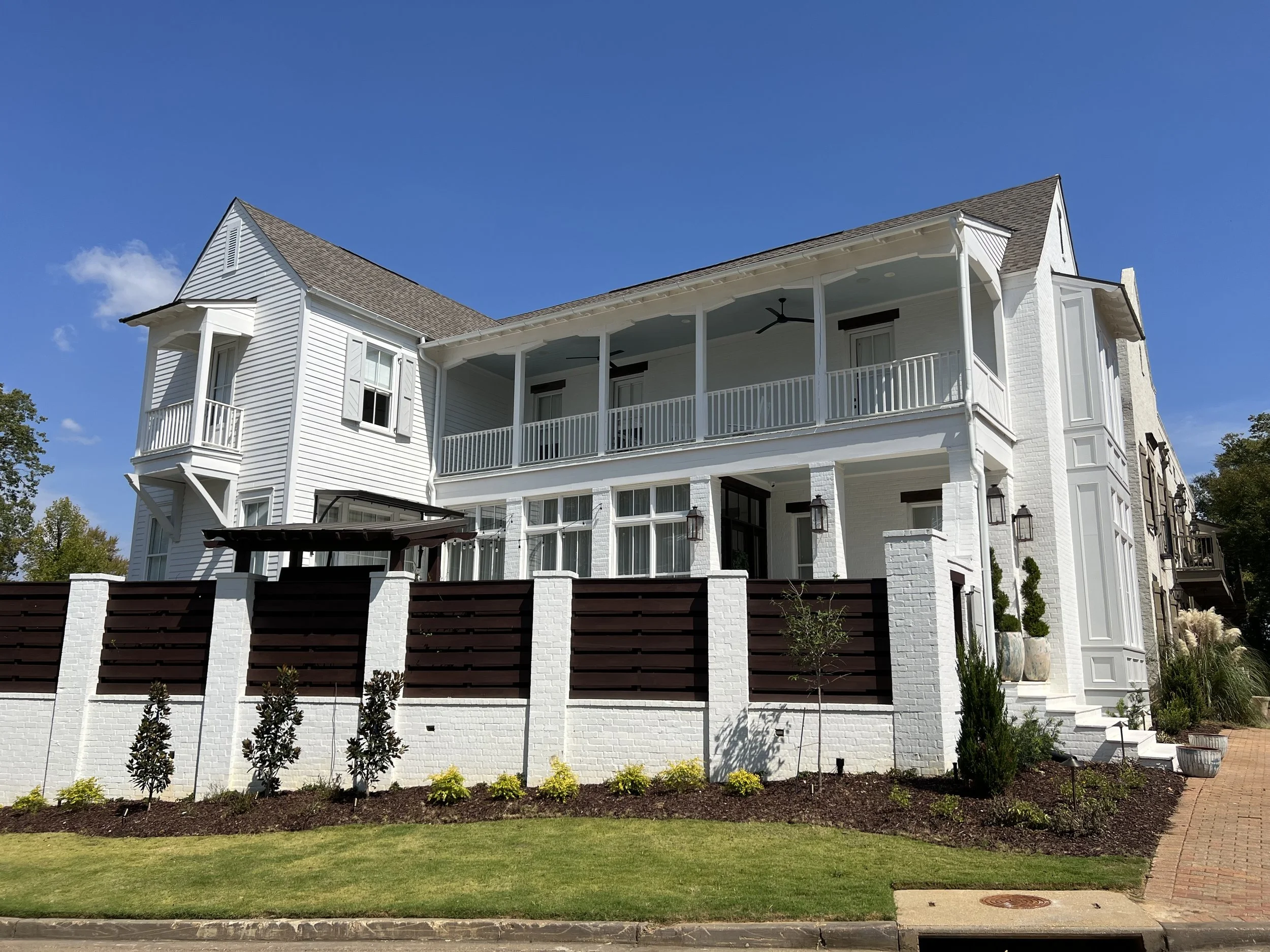 Large white multi-story house with a landscaped yard, brick fence with dark wood accents, and a brick walkway, under a clear blue sky.