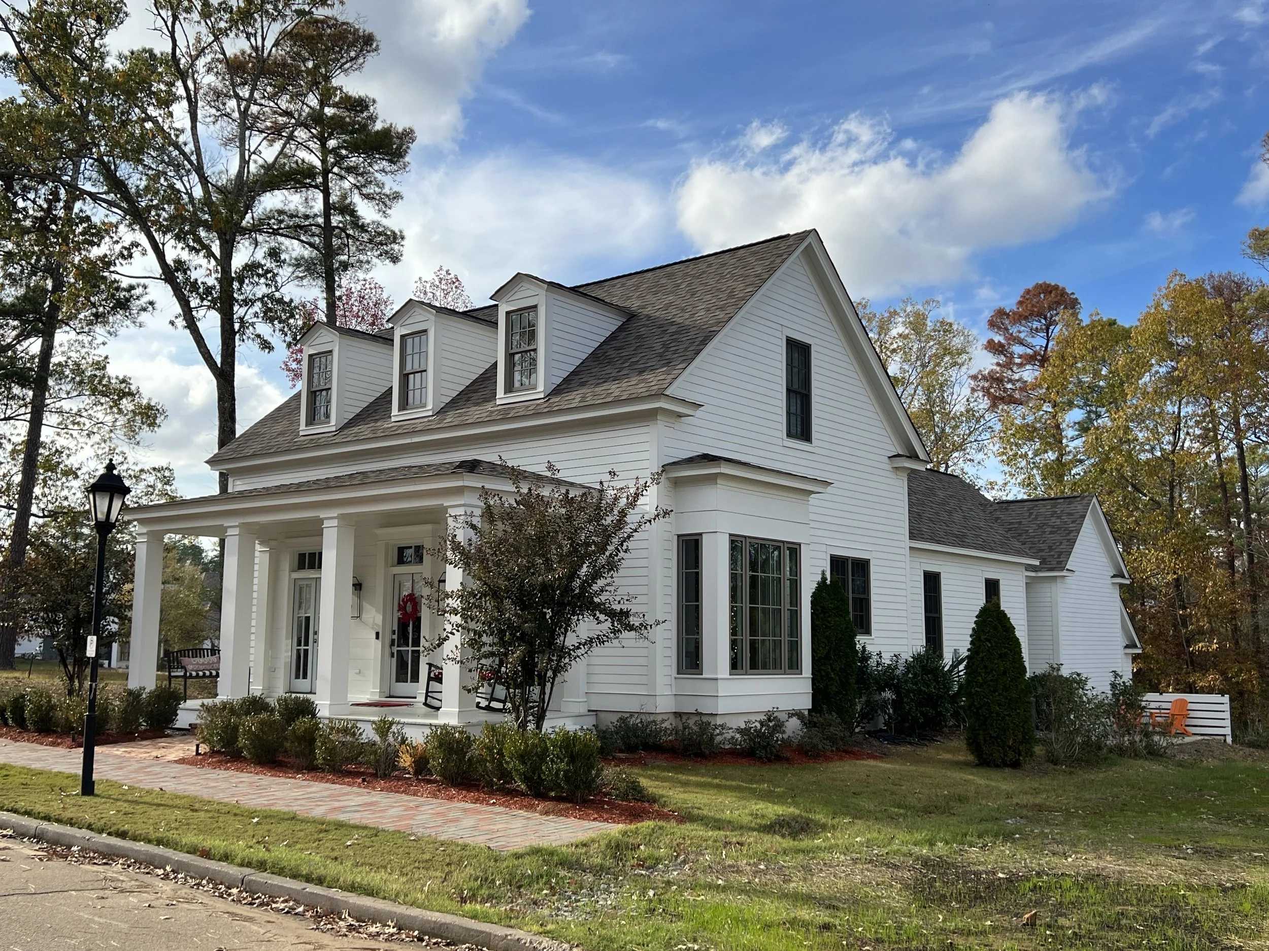 A large white house with a gabled roof, dormer windows, and a front porch, surrounded by trees and a lawn with a sidewalk and lamppost in front.