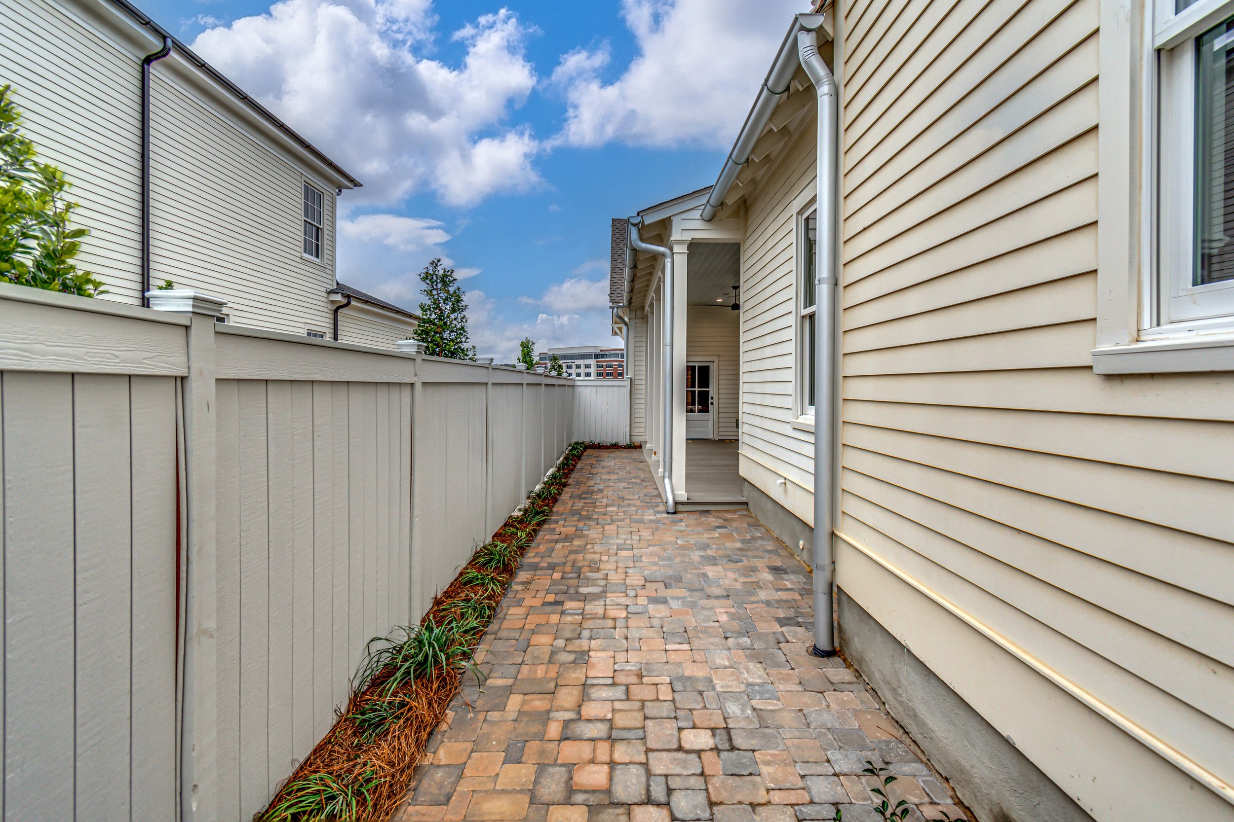 Backyard with brick pathway, beige house siding, white fence, and a small garden bed with plants, under a partly cloudy sky.