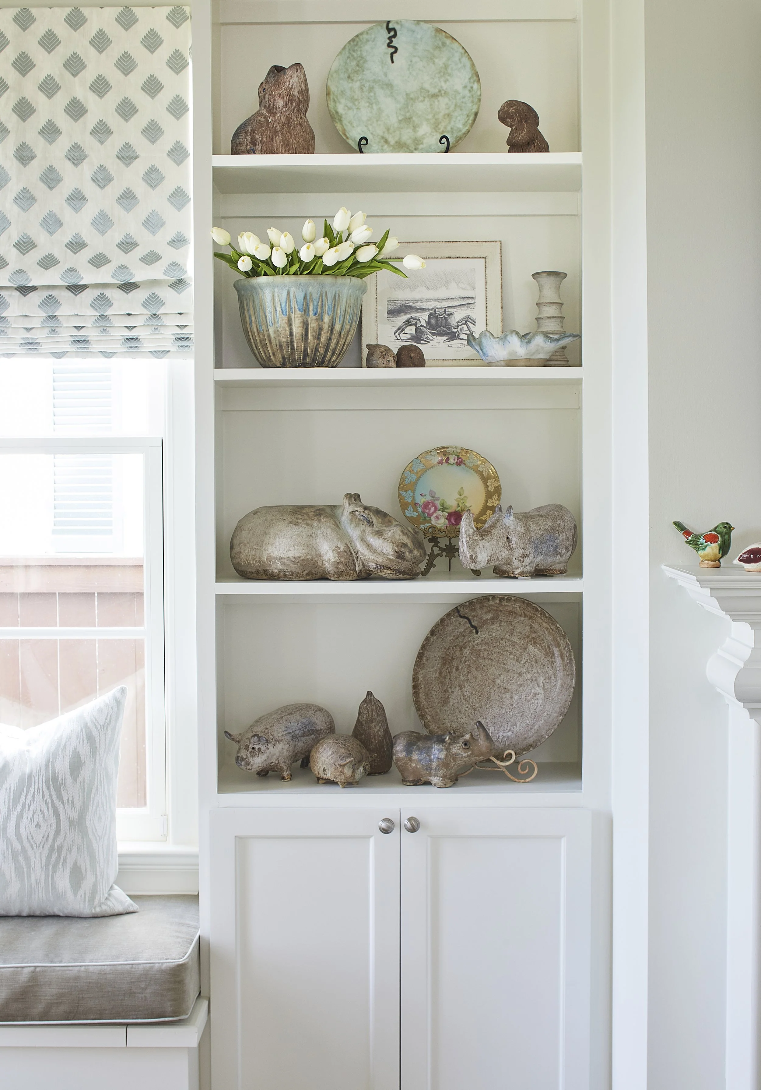 A built-in white bookshelf with decorative animal-shaped pottery, plates, and a vase with white tulips, situated next to a window with patterned Roman shade.