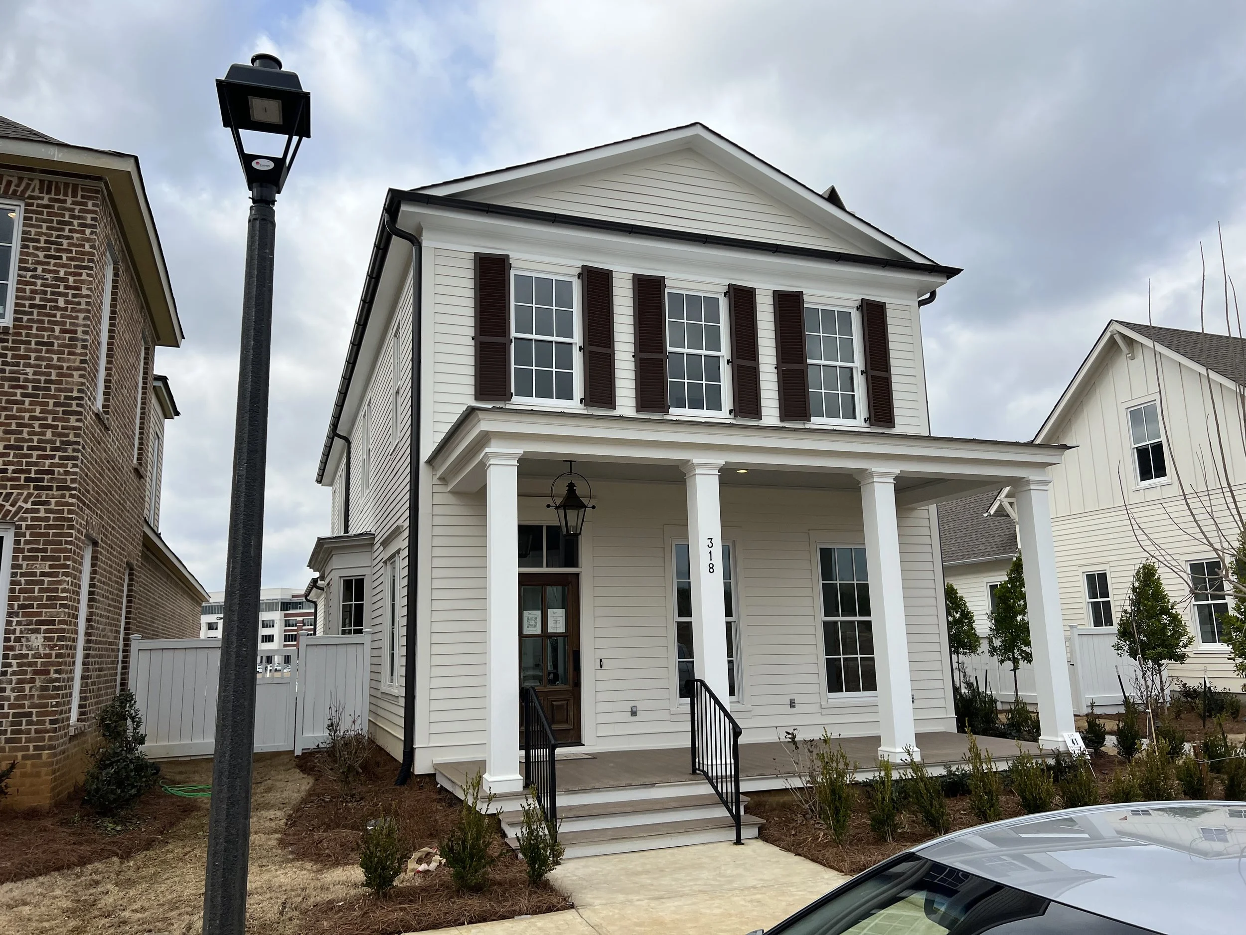 White three-story house with dark shutters, front porch with columns, and a black lamp hanging near the door. A street lamp is visible in the foreground, and neighboring houses are on either side.