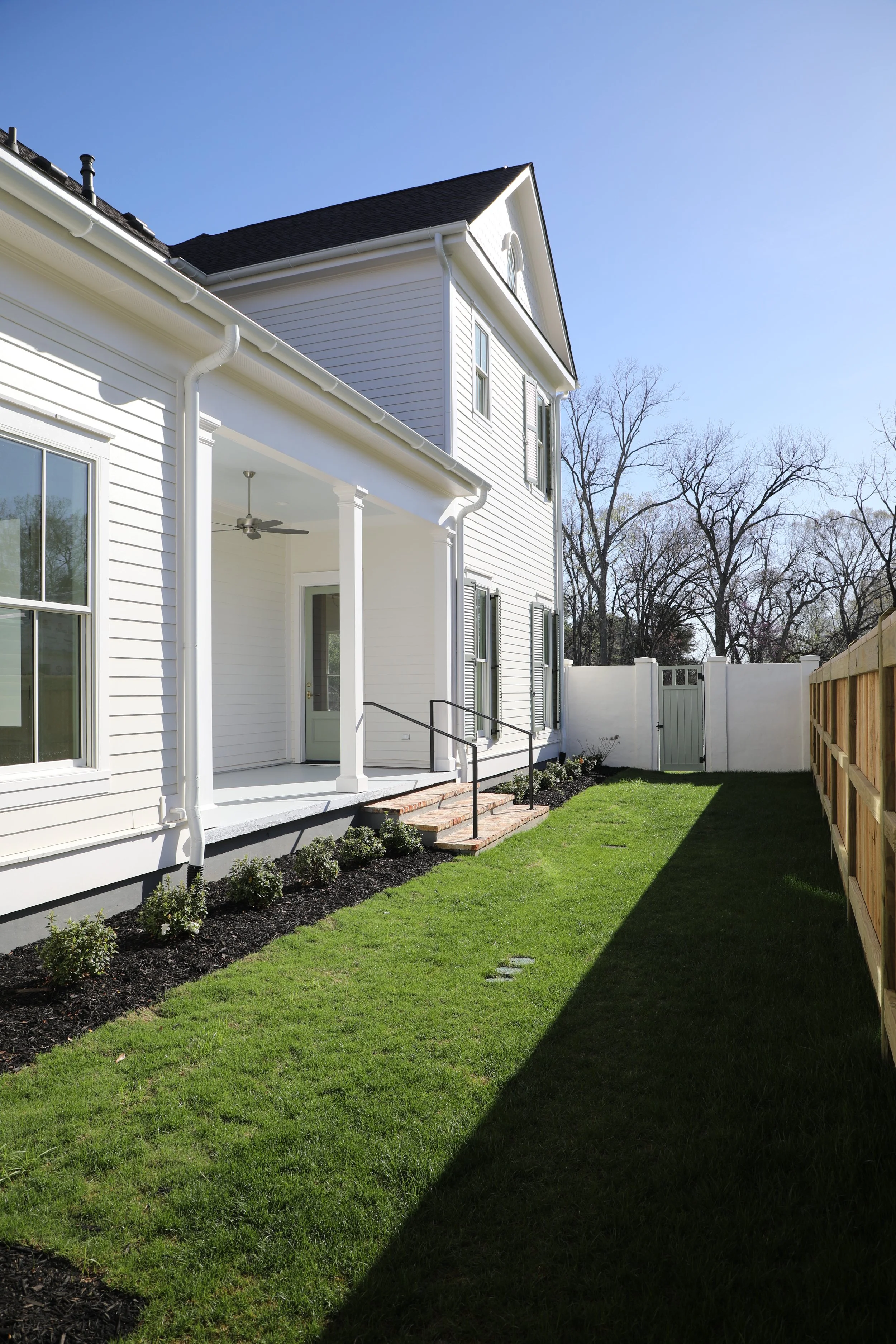 Backyard with a small porch, green lawn, and a white house with black roof, white fence, and wooden fence against a clear blue sky.