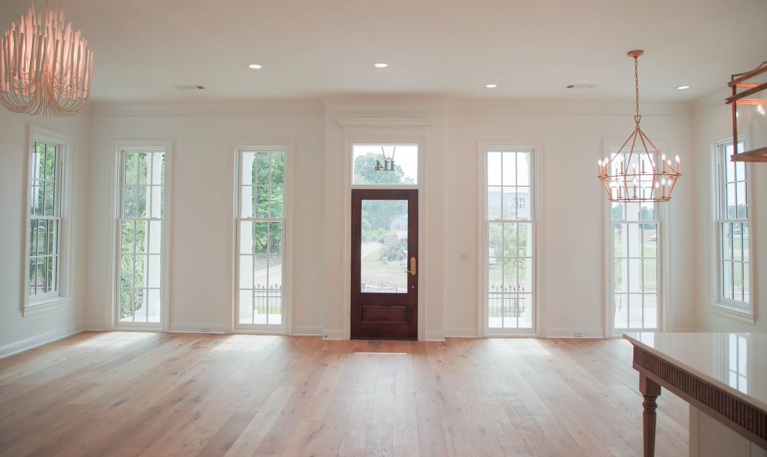 Empty room with hardwood floors, white walls, large windows, and a central wooden door, natural light, and chandeliers on the ceiling.